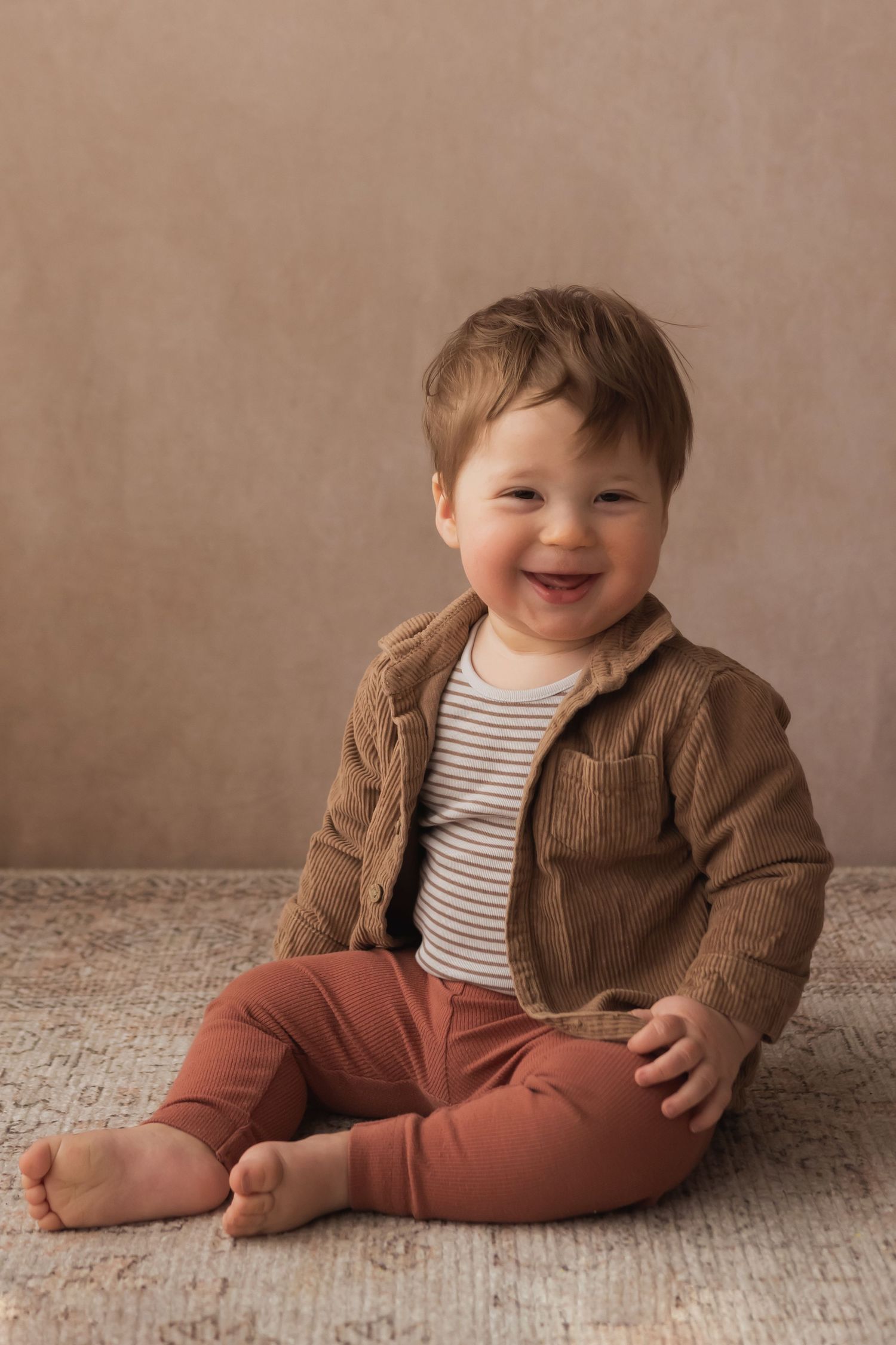 Adorable laughing baby boy in brown corduroy jacket and rust pants sits barefoot on a soft rug.