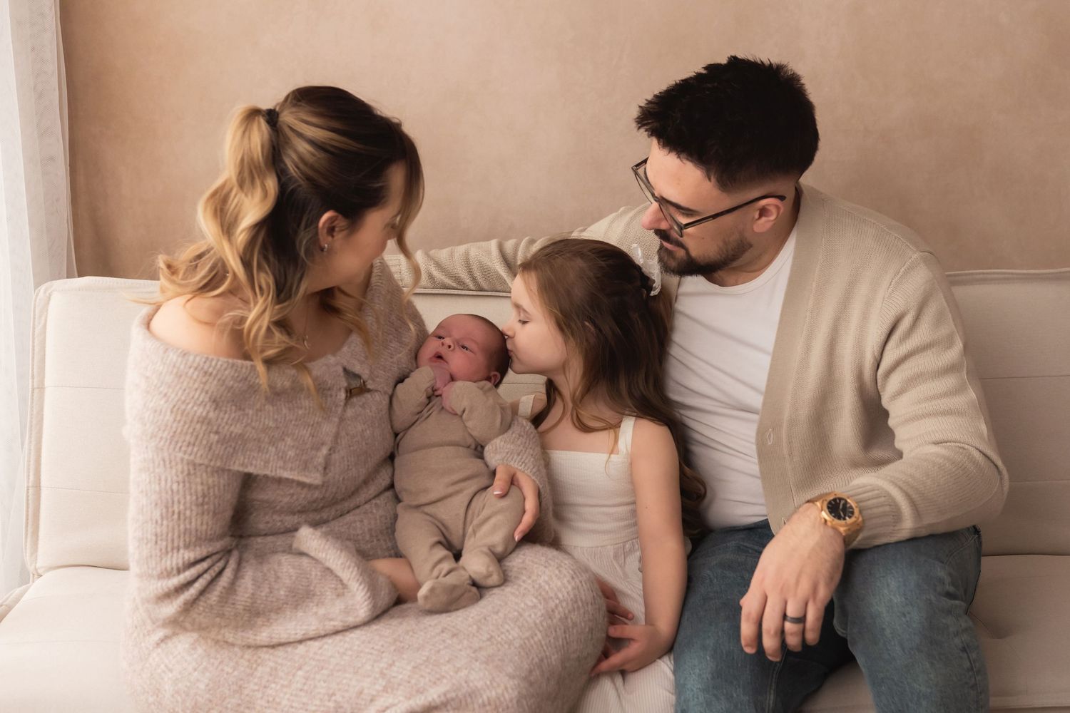 Family of four sitting on a couch, parents lovingly gazing at newborn baby held by mother, with young daughter nearby.