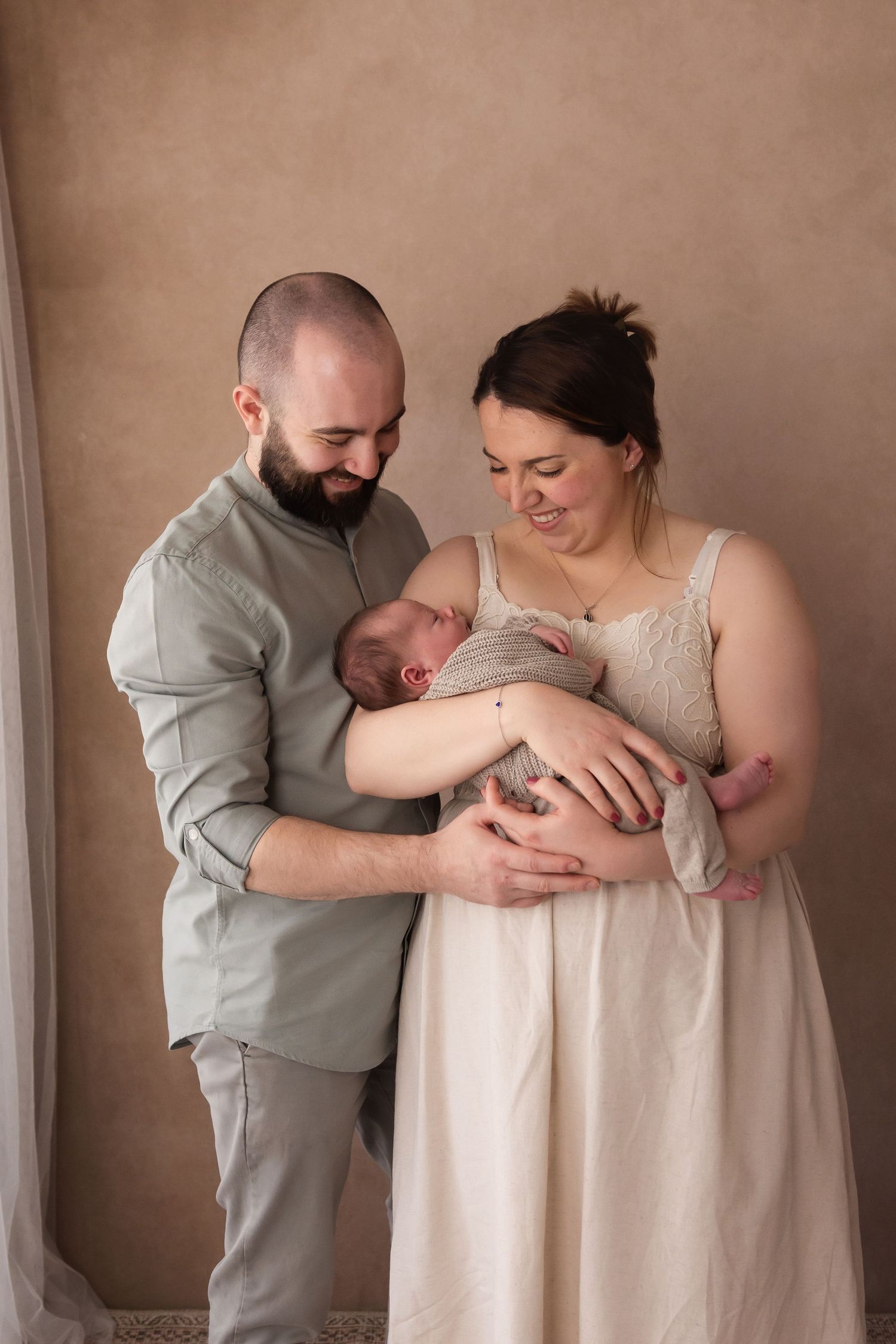 New parents standing together holding their newborn baby against a warm beige backdrop.