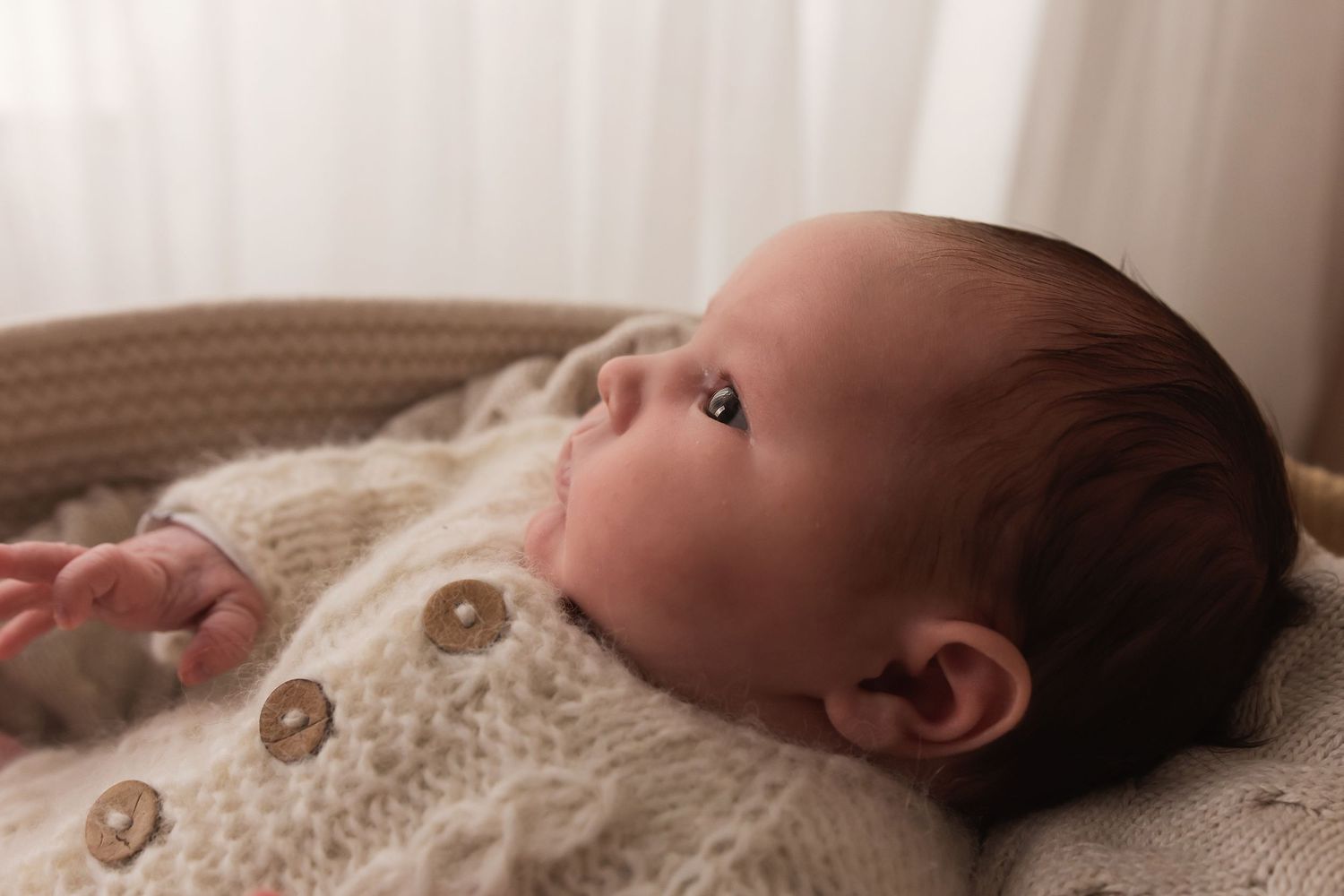Close-up of newborn baby with dark hair in cream knit cardigan lying in basket.
