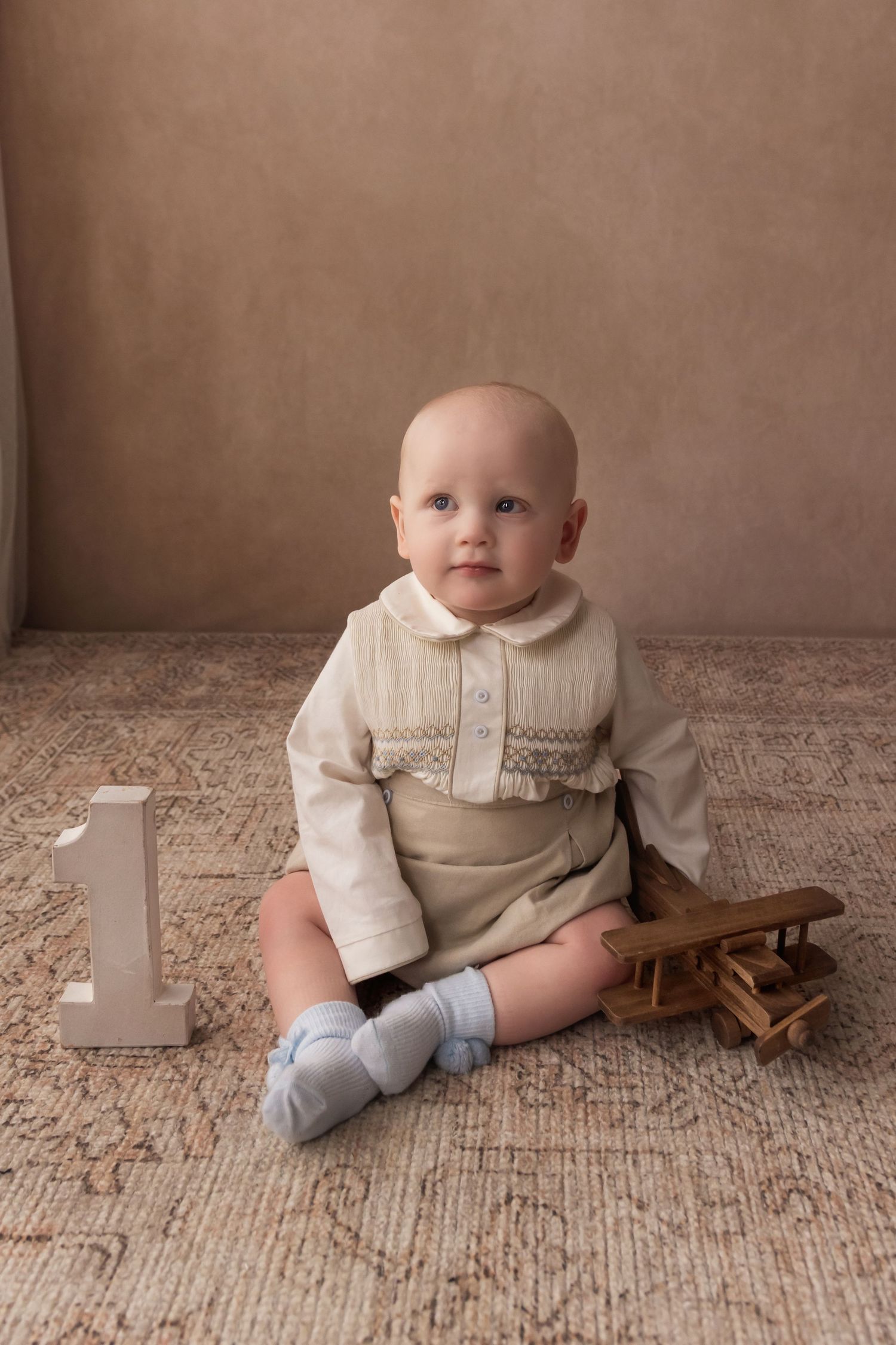 Sweet baby boy in smocked cream outfit sits with wooden toy airplane and number 1 block.