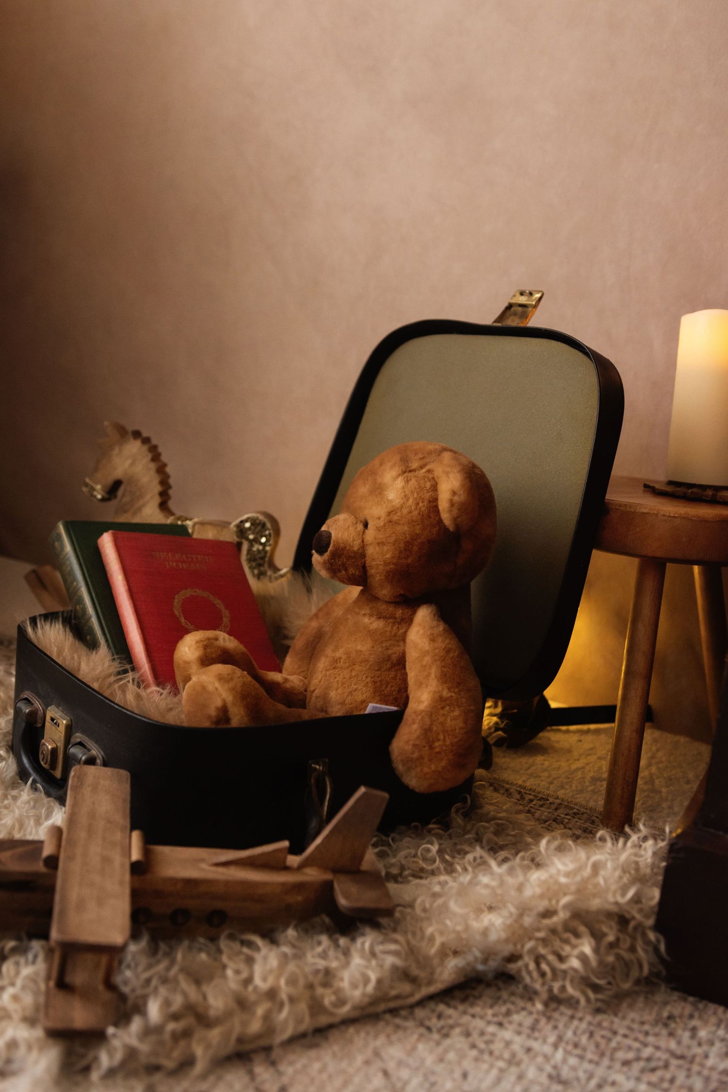 Vintage teddy bear sitting in an open suitcase with books, beside a glowing candle on a wooden stool.