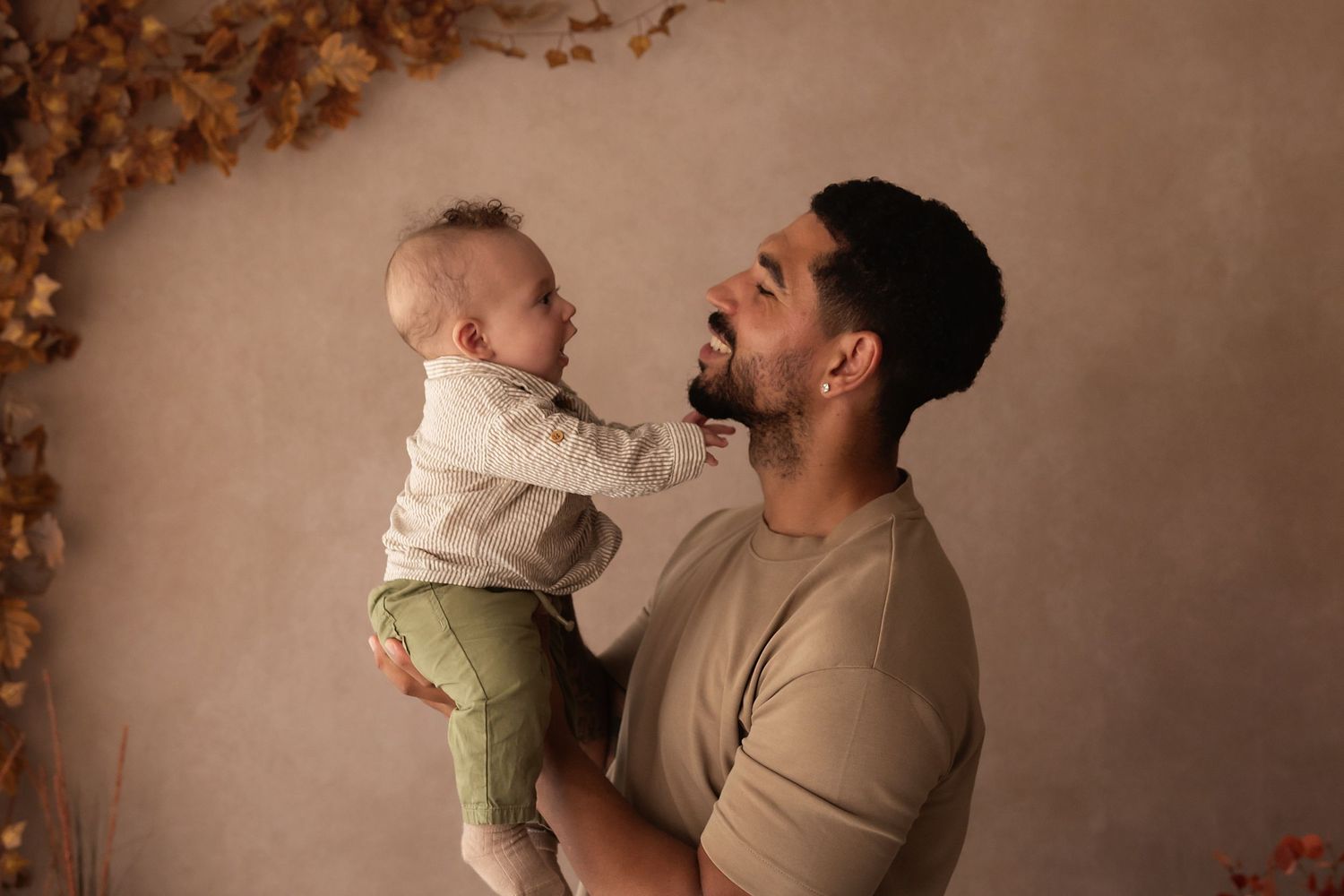 A bearded man lovingly holds a smiling baby in green pants, sharing a joyful moment against a warm studio backdrop.