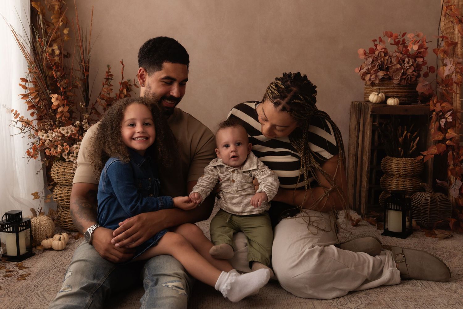 A happy mixed-race family of four poses together on a floor with autumn decor, including a baby and young girl.