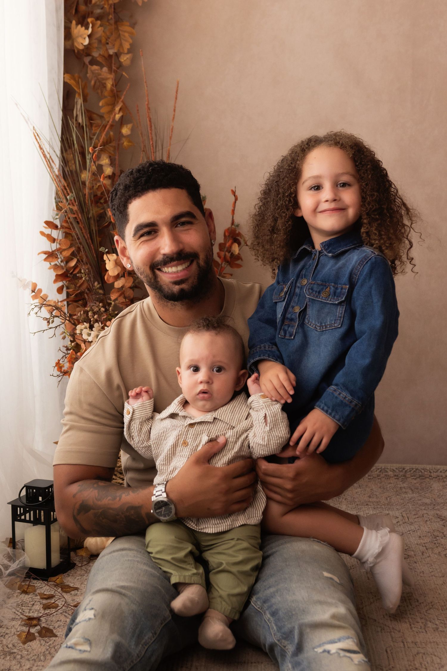 A smiling father sits with his baby and young curly-haired girl in a warm autumn-themed studio portrait.