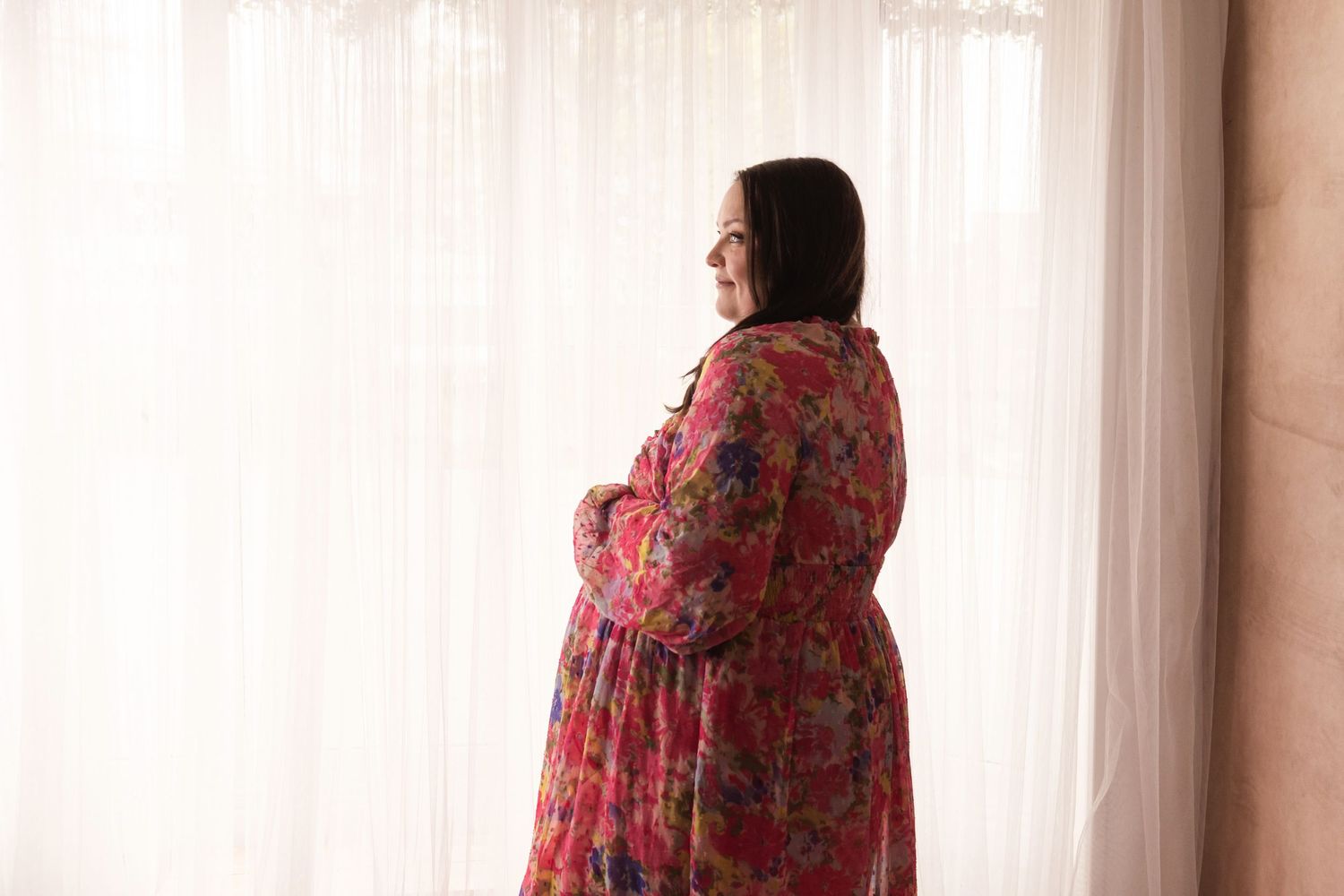 Pregnant woman in colorful floral dress standing by bright sheer curtains, smiling sideways.