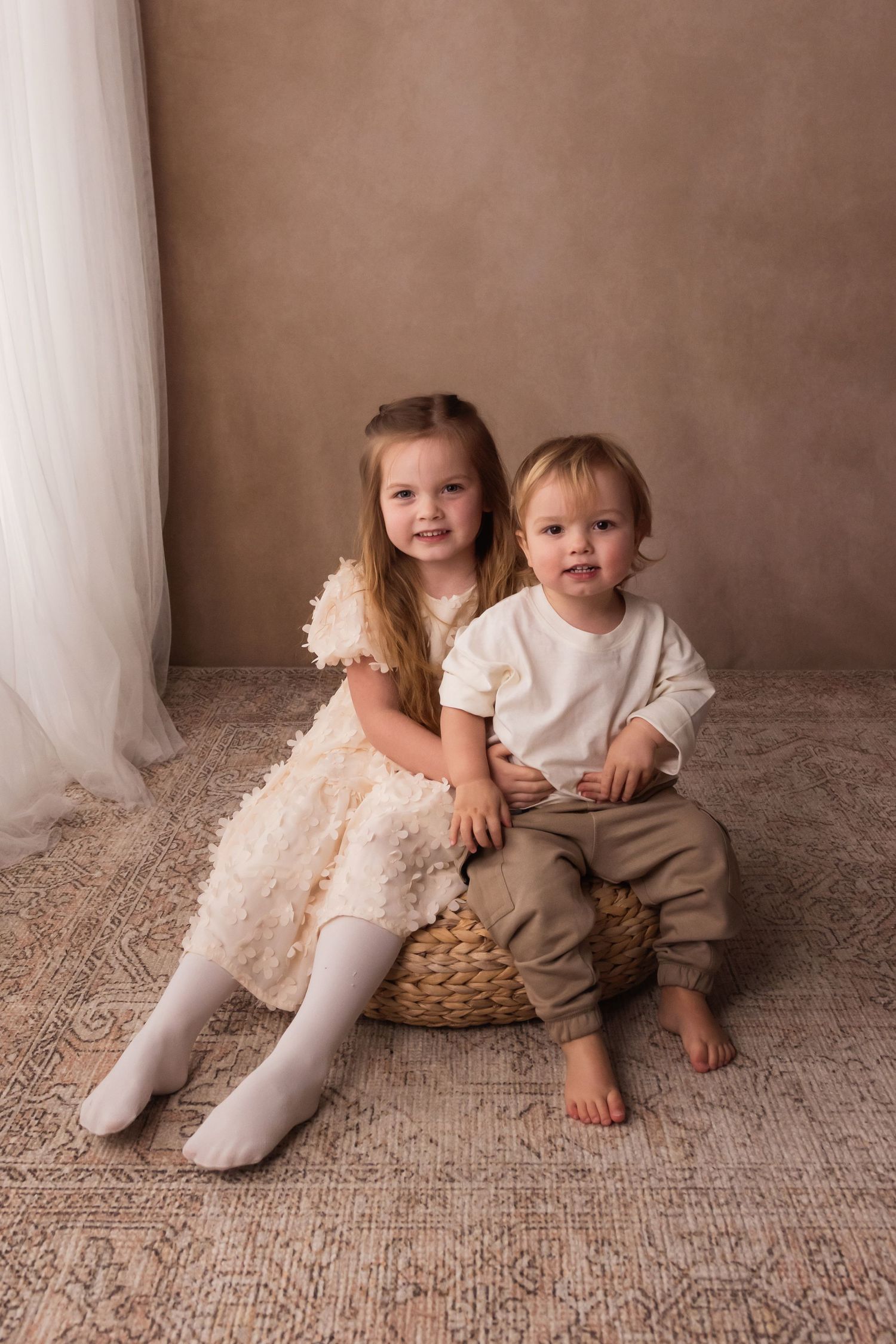 Two young siblings posing together on a wicker stool during a professional studio photo session.
