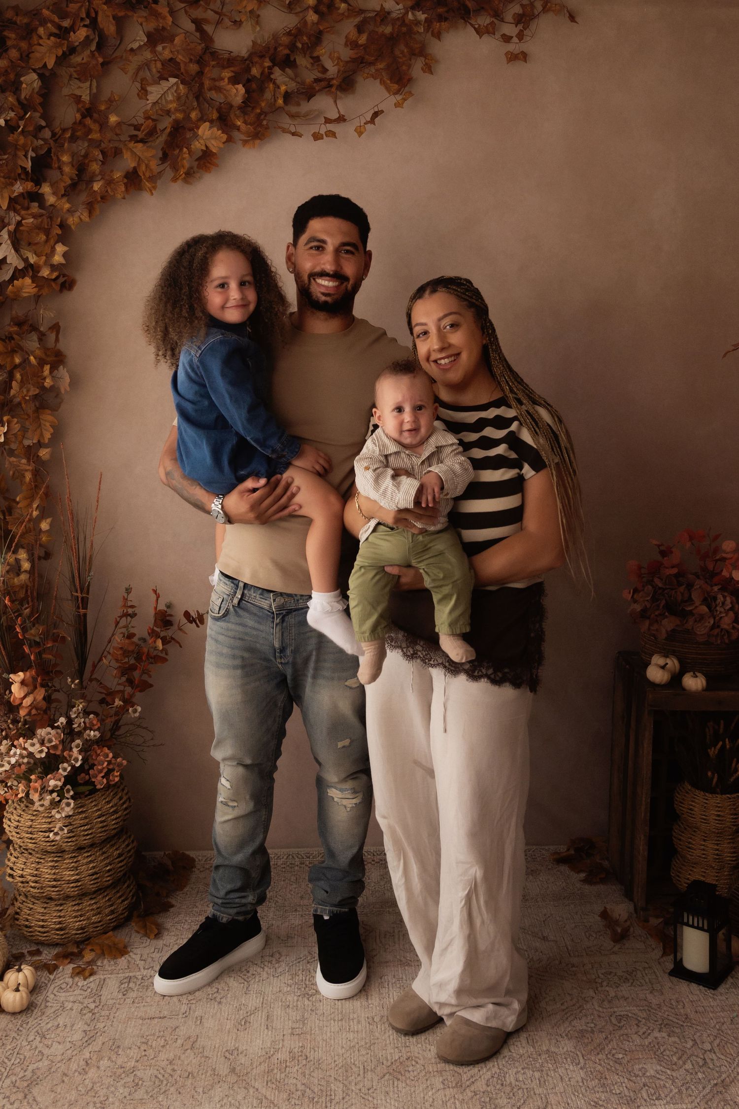 A smiling family of four poses for a fall portrait, with parents holding two young children against a warm autumn backdrop.