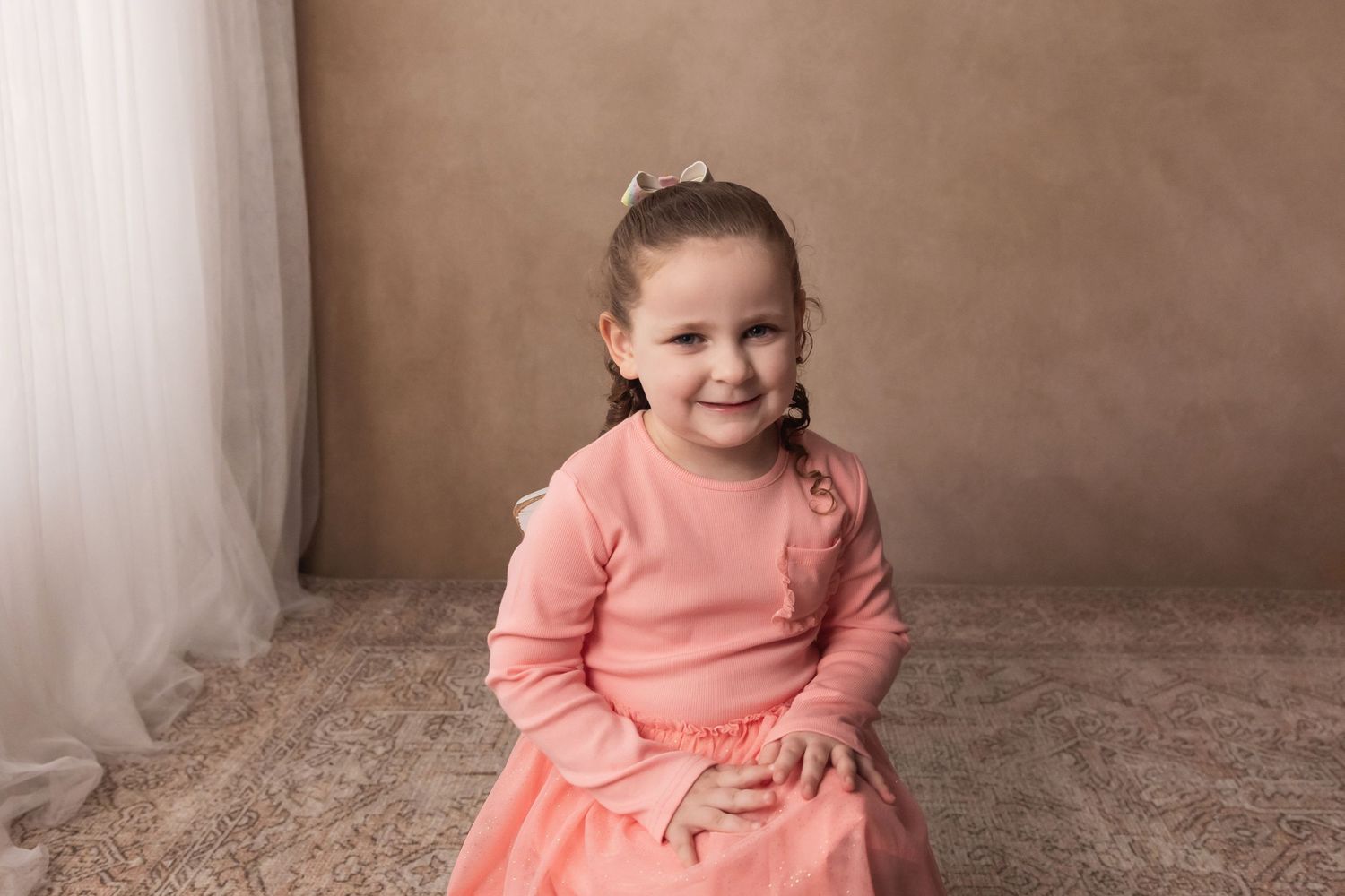 A smiling young girl in a pink dress sits on a rug in a professional photo studio portrait.
