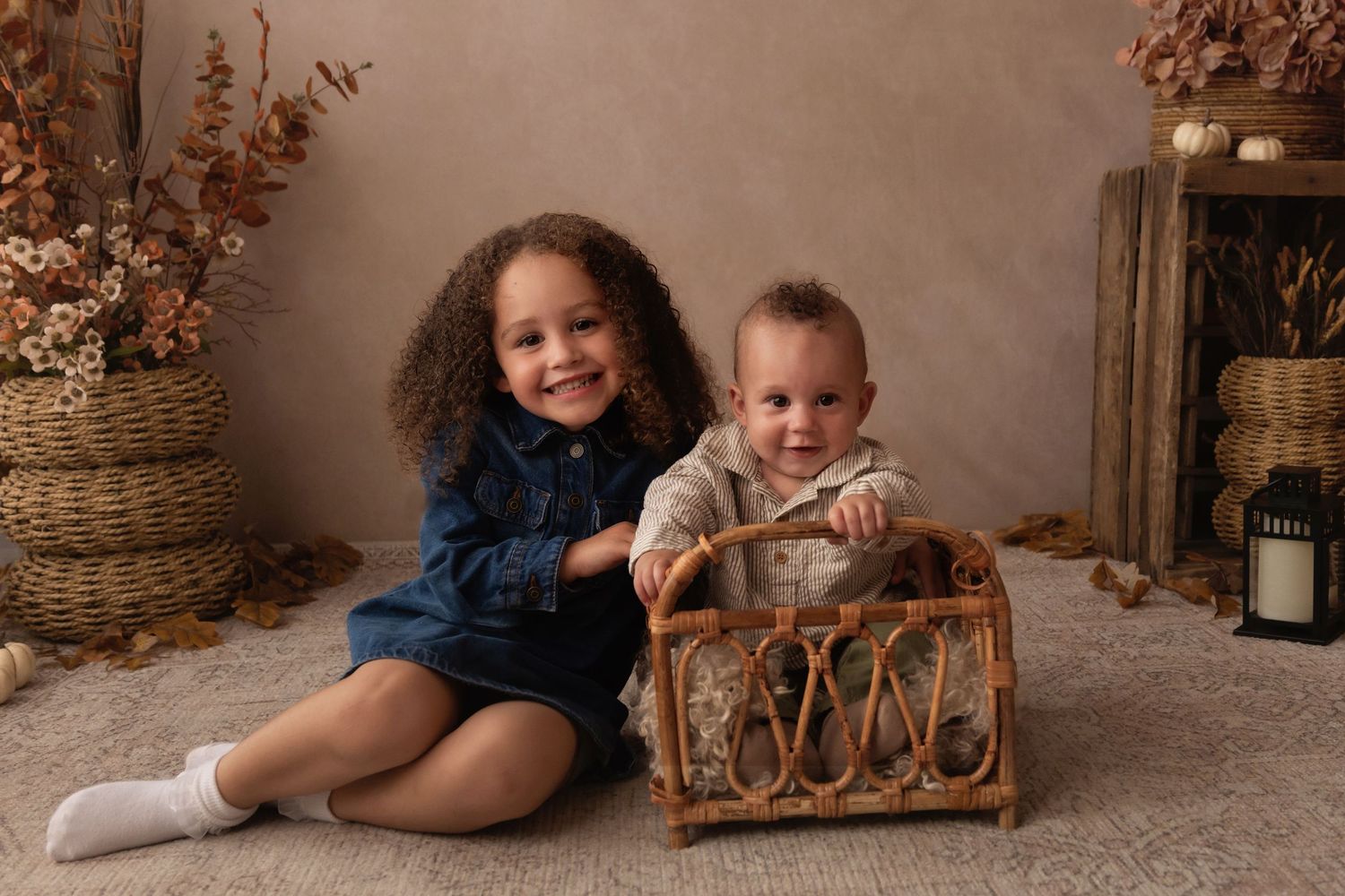 Two young children pose with a wicker basket in a cozy fall-themed studio portrait with warm autumn decor.