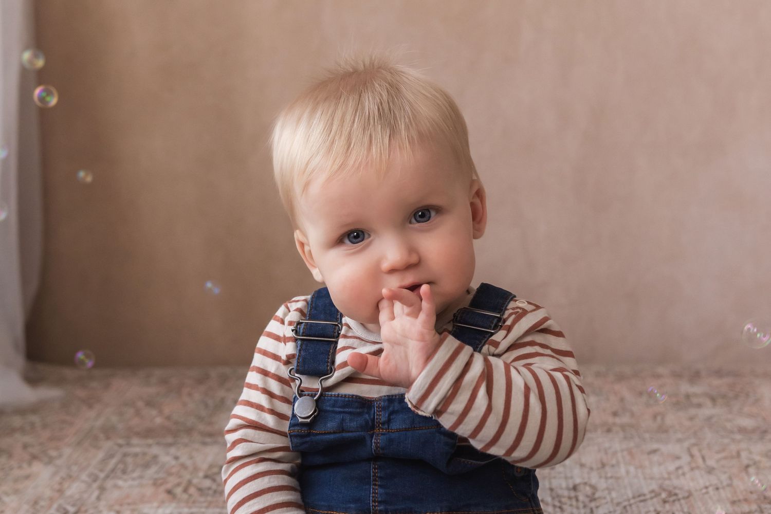 An adorable blonde baby in denim overalls sits on a rug with finger in mouth, surrounded by bubbles.