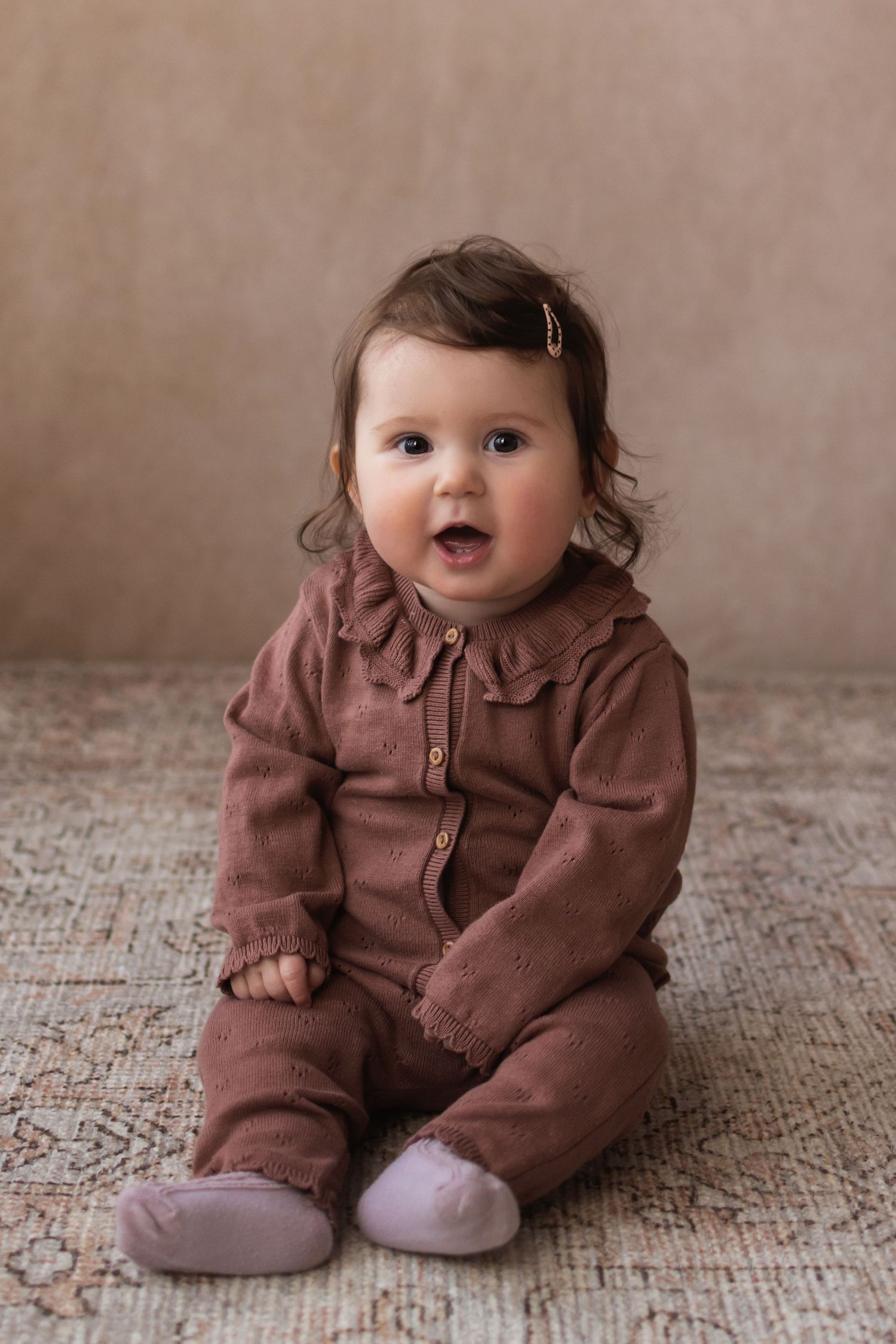 A cheerful baby girl in a brown romper sits on a rug smiling brightly in a studio portrait.