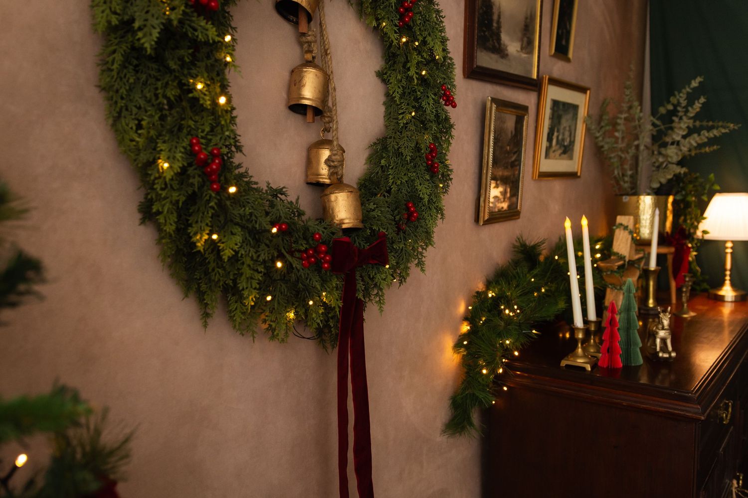 A festive Christmas wreath with gold bells, red berries, and warm lights hangs on a wall beside holiday garland and candles.