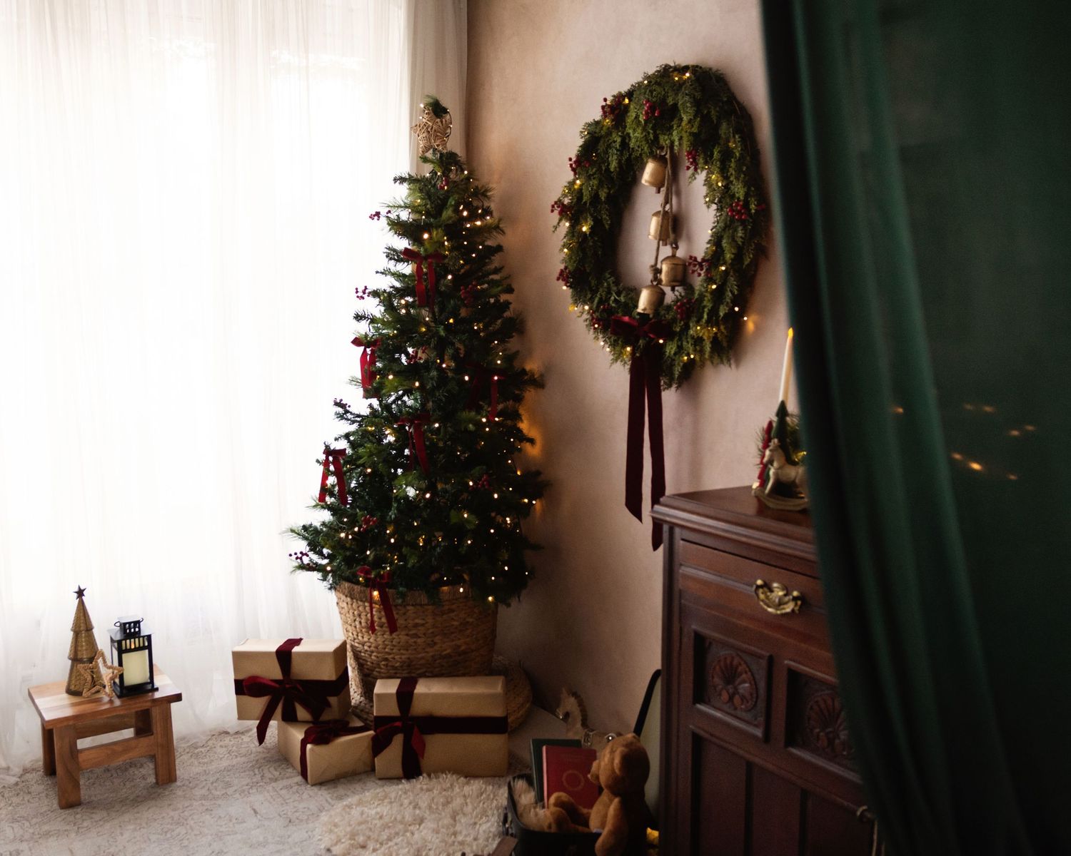 Cozy Christmas corner with decorated tree, gift boxes, wreath on wall, and wooden dresser with green curtain.