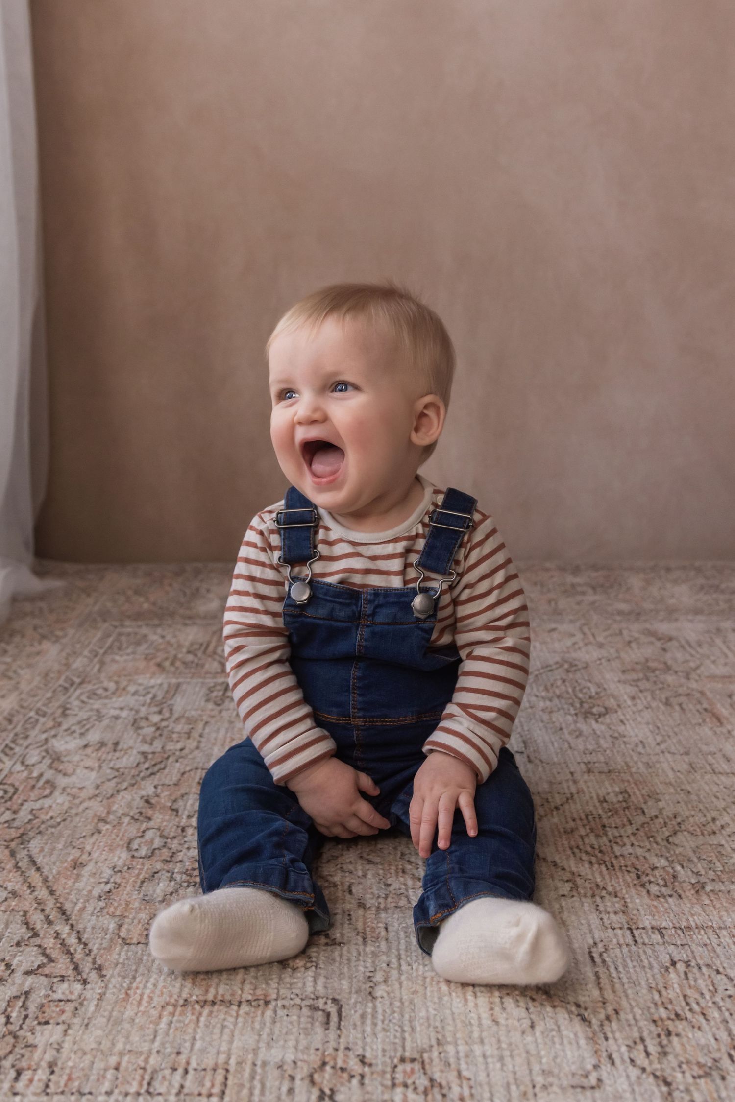 A happy blonde baby in denim overalls sits on a patterned rug, laughing with mouth wide open.