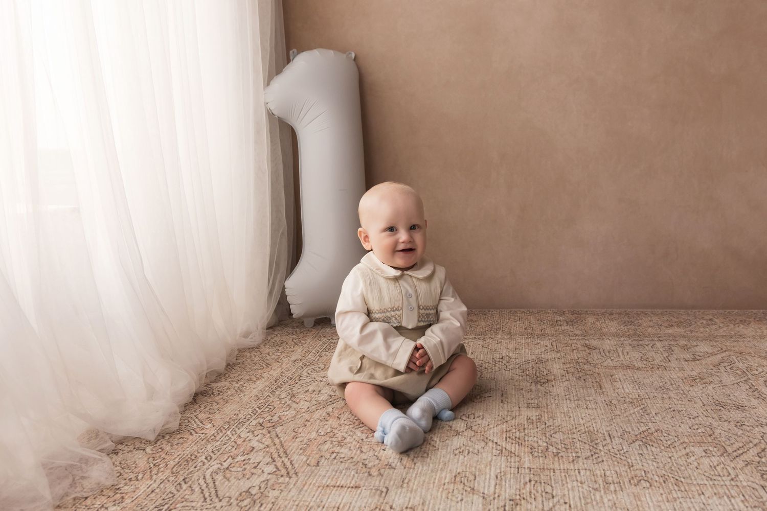Smiling baby boy in beige outfit sits on rug beside number 1 balloon for first birthday.