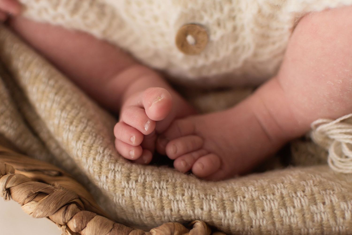 Close-up of tiny newborn baby feet resting in wicker basket with cream knit outfit.