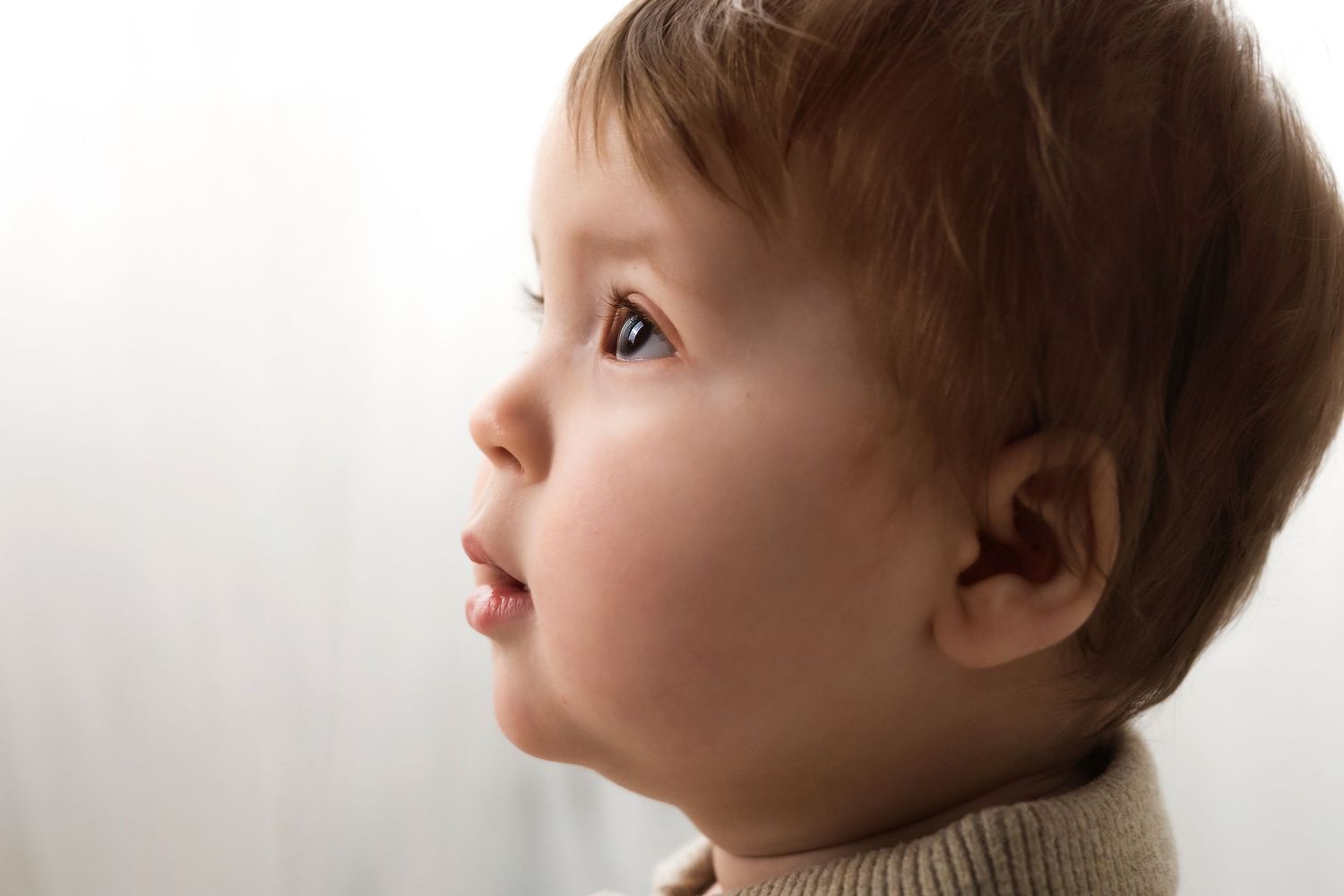 Close-up profile of a curious toddler with brown hair and blue eyes looking upward in soft natural light.