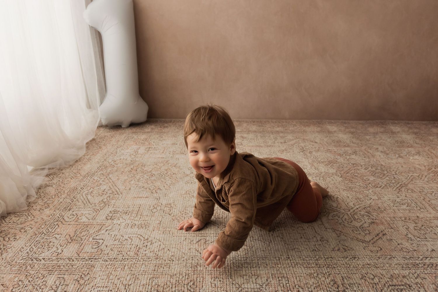 Happy crawling baby in brown outfit smiling on a patterned rug near white curtains.