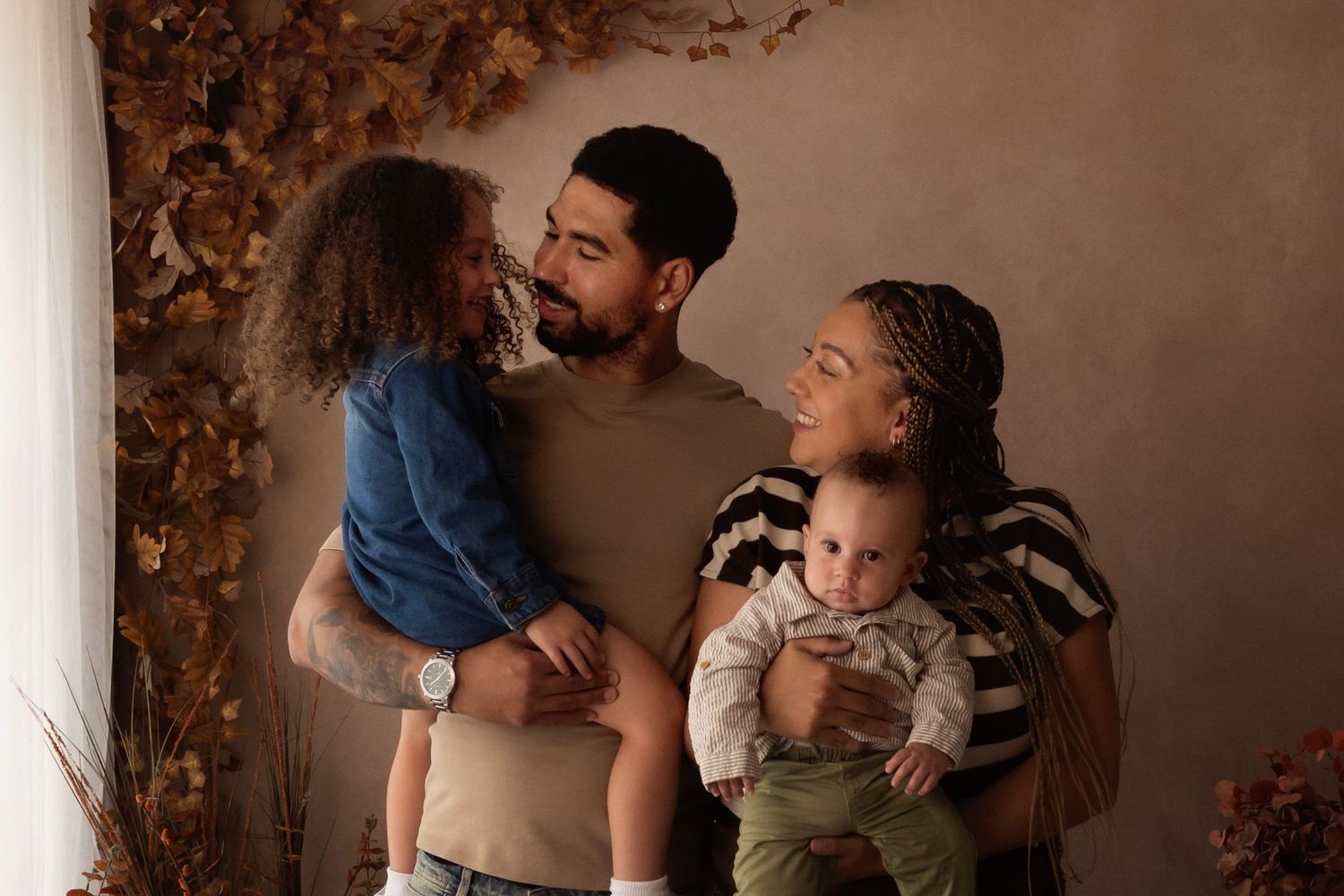 A loving family of four poses together, parents holding two young children against a warm brown backdrop with autumn leaves.