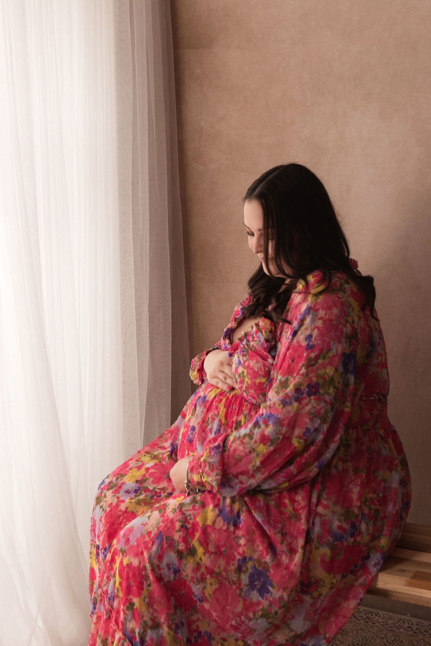 Pregnant woman in a vibrant floral dress gently cradling her baby bump while sitting by a window.
