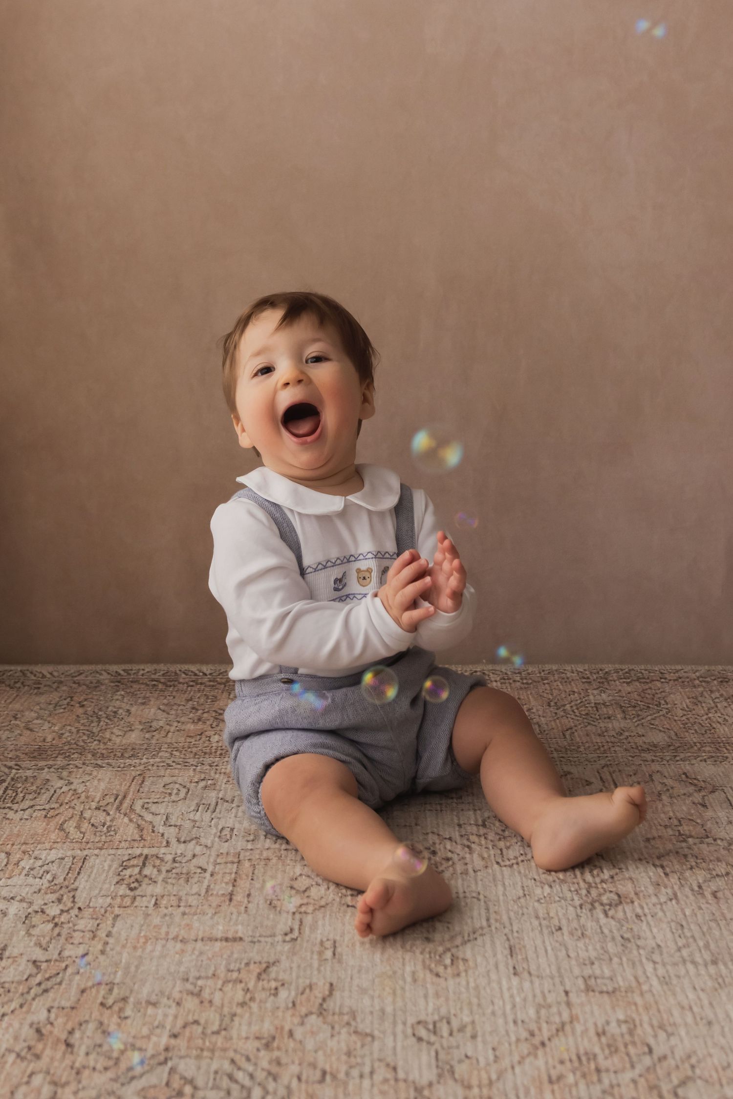 Laughing baby boy in white shirt and blue shorts sitting on rug reaching for soap bubbles.