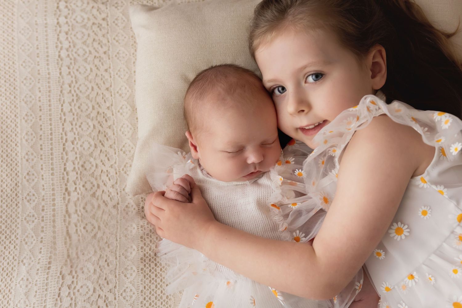 Big sister in daisy dress cuddling sleeping newborn baby on cream knit blanket.