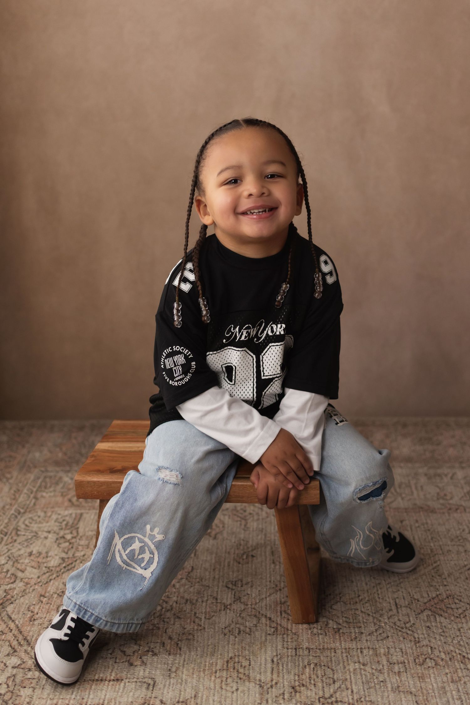 A cheerful young child with braids in a New York jersey and ripped jeans sits on a wooden stool smiling.