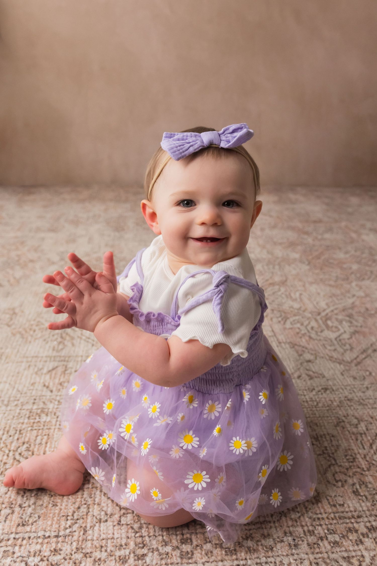 Joyful baby girl in purple daisy dress clapping hands and smiling during birthday photo session.