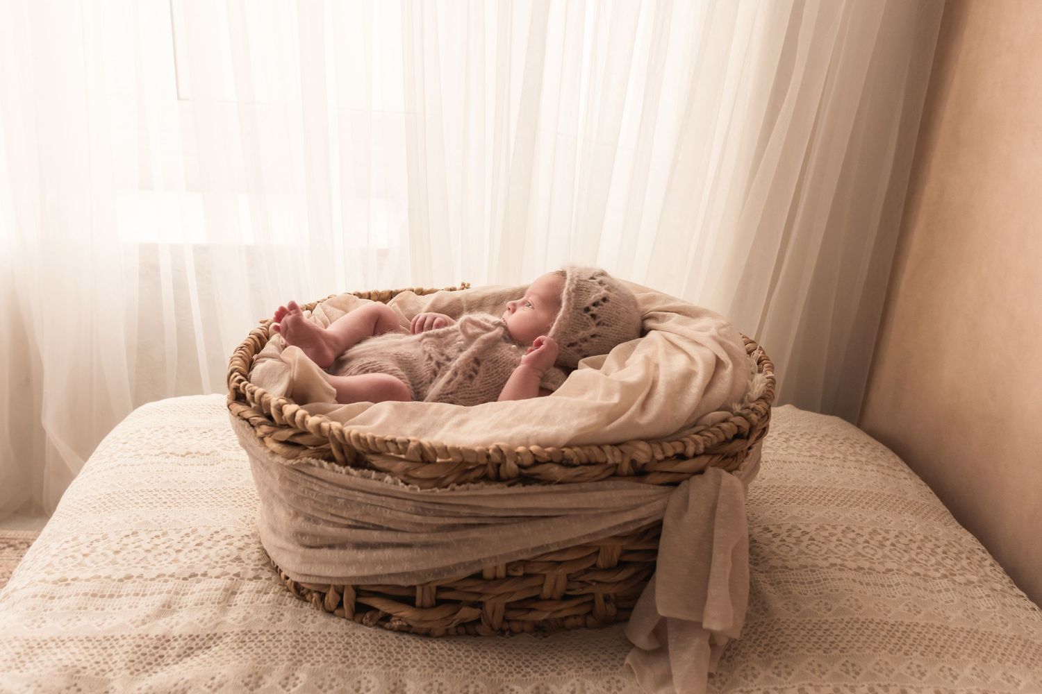Newborn baby sleeping in wicker basket with pink wrap near sheer white curtains.