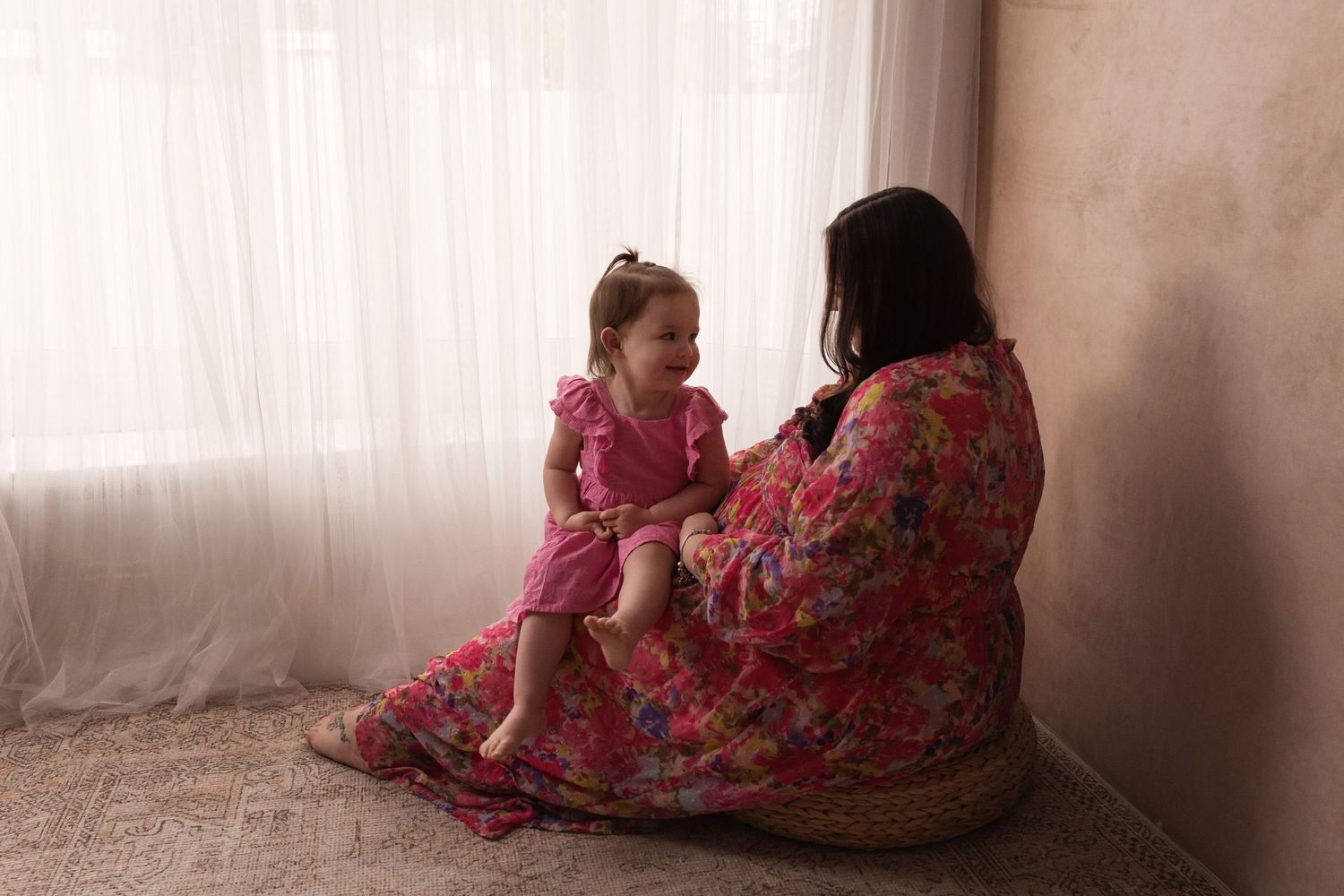 Pregnant woman in floral dress sitting on floor with toddler girl near sheer curtains.