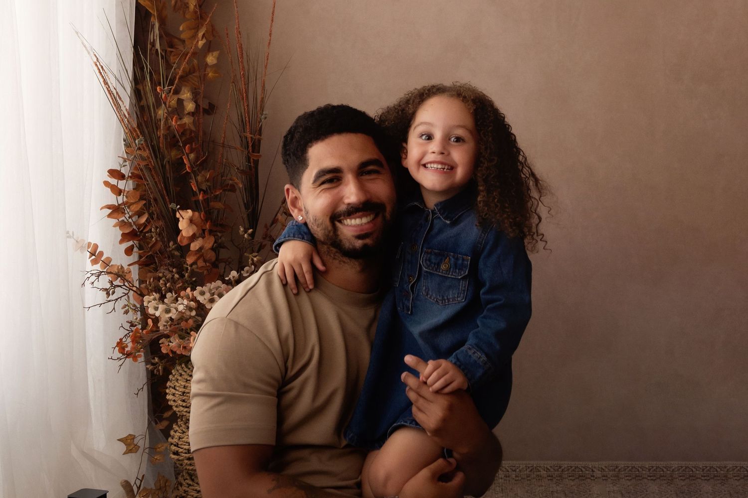 A smiling man holds a happy curly-haired girl in a denim shirt, posing before dried floral decor.