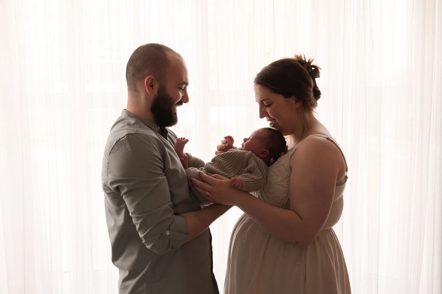 New parents standing by white curtains tenderly holding their newborn baby together.