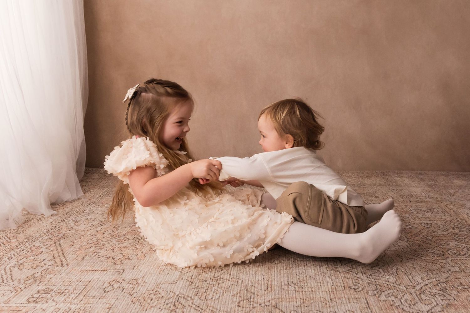 Two young children in formal attire sit on a rug, smiling and holding hands in a warm studio setting.