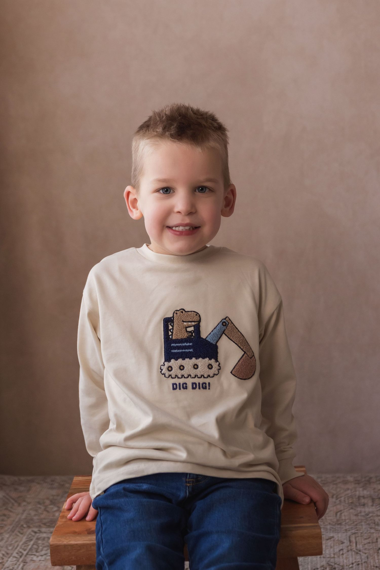 A smiling young boy sits on a wooden bench wearing a beige long-sleeve shirt with a digger print.
