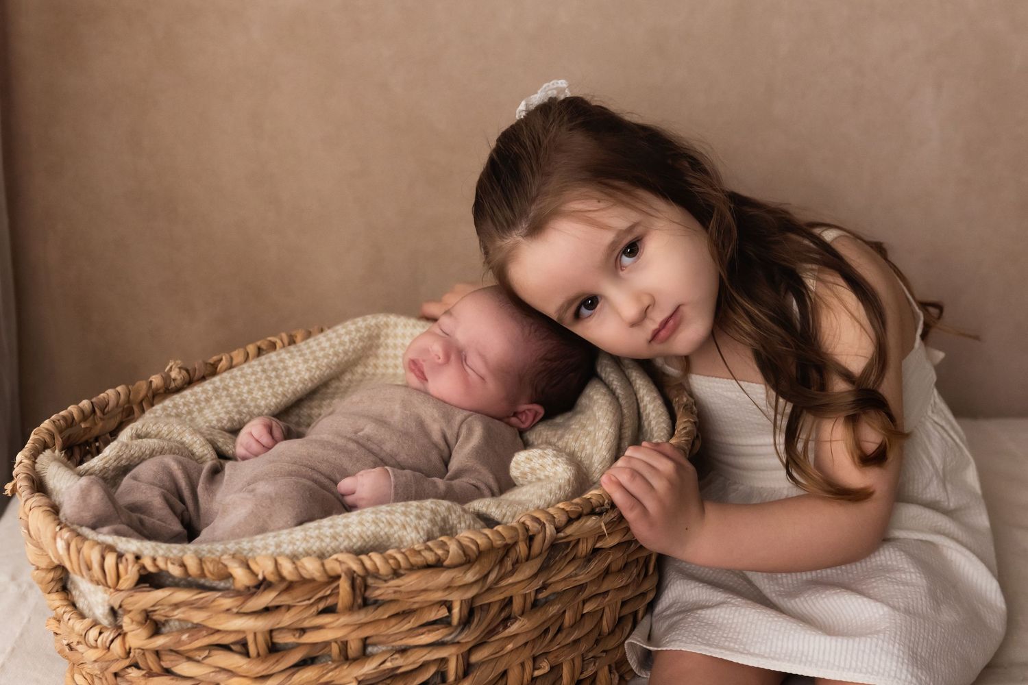Young girl with brown hair posing proudly beside sleeping newborn sibling in wicker basket.