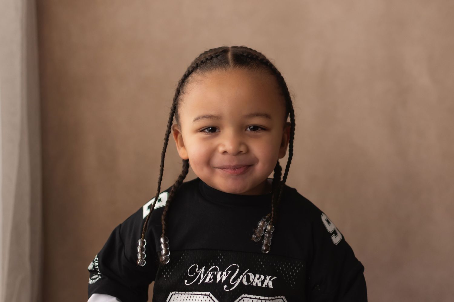 Smiling young child with braids wearing a black New York jersey poses for a portrait photo.