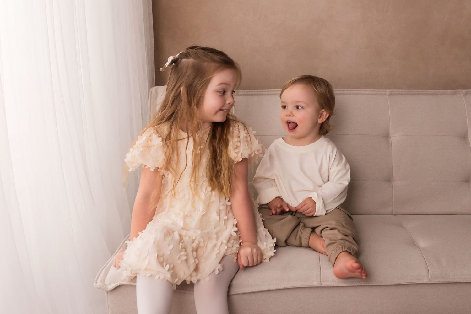 A little girl in a cream ruffled dress sits on a sofa smiling at a laughing baby beside her.