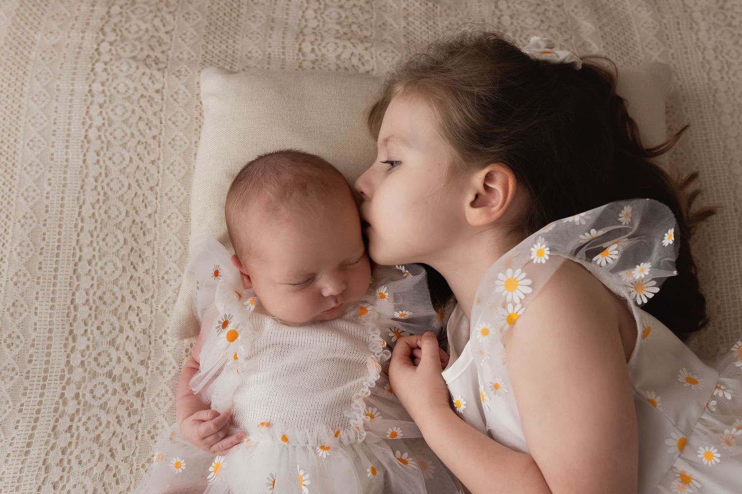Older sister gently kissing sleeping newborn baby lying on cream blanket.