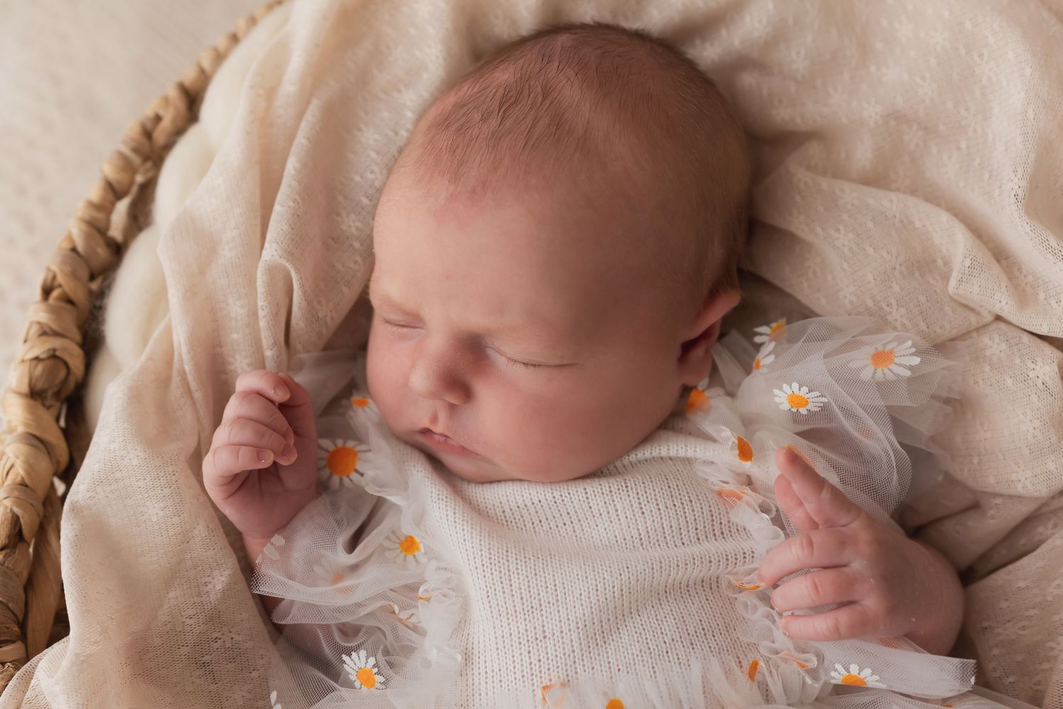 Close-up of sleeping newborn baby in white daisy dress resting in wicker basket.