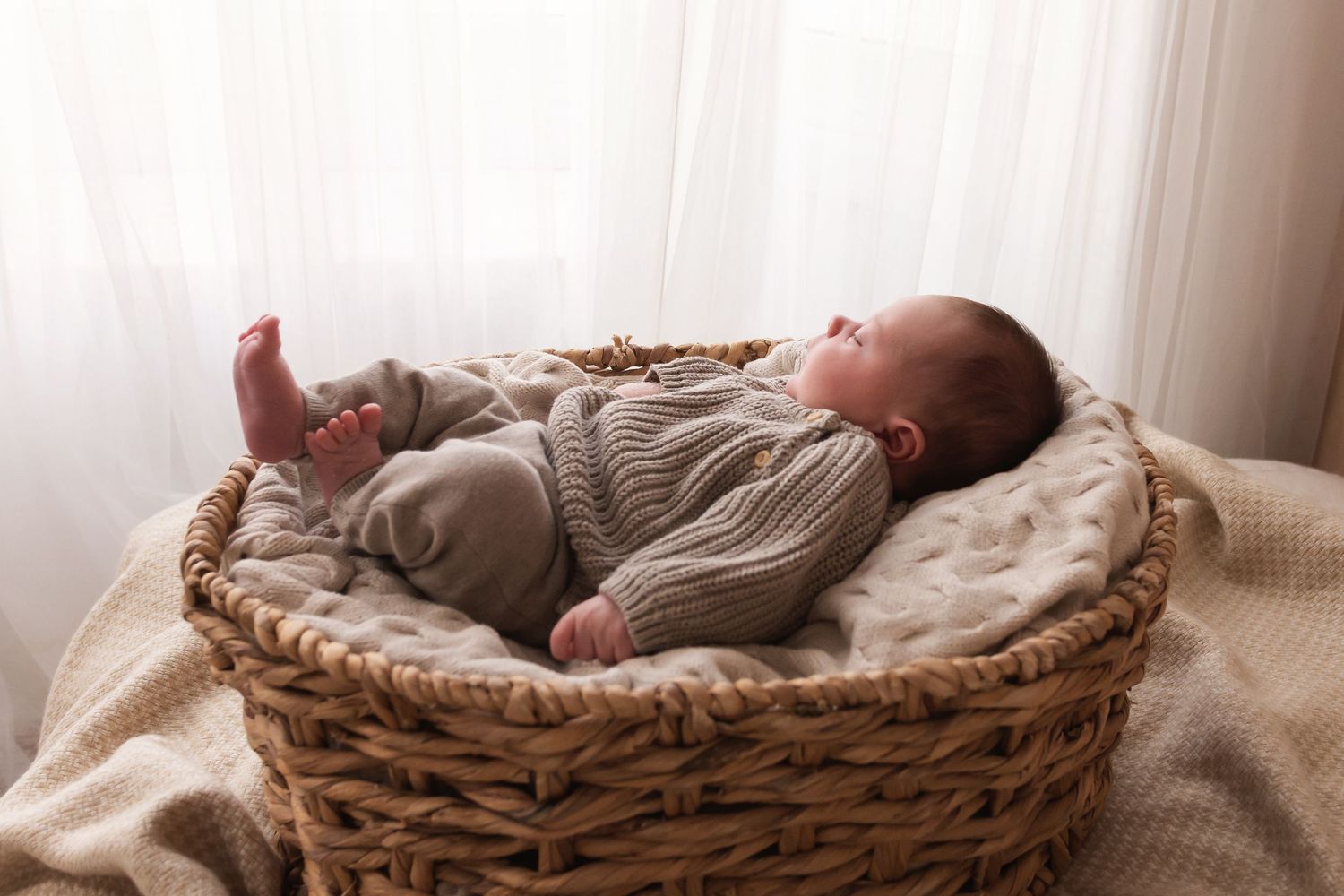 Newborn baby in knit outfit lying in wicker basket with soft blanket near window.