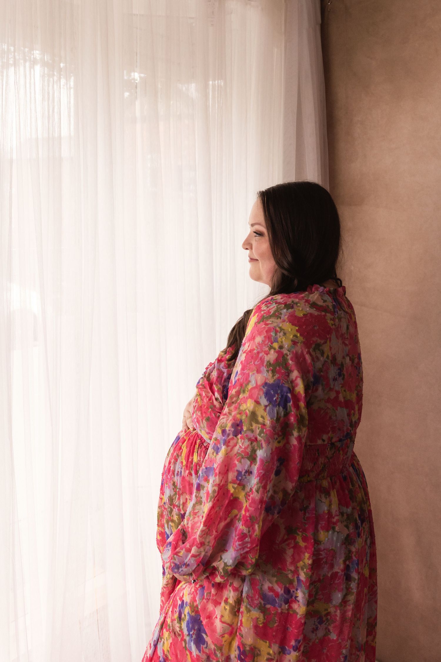 Side profile of pregnant woman in colorful floral dress leaning against wall near white curtains.