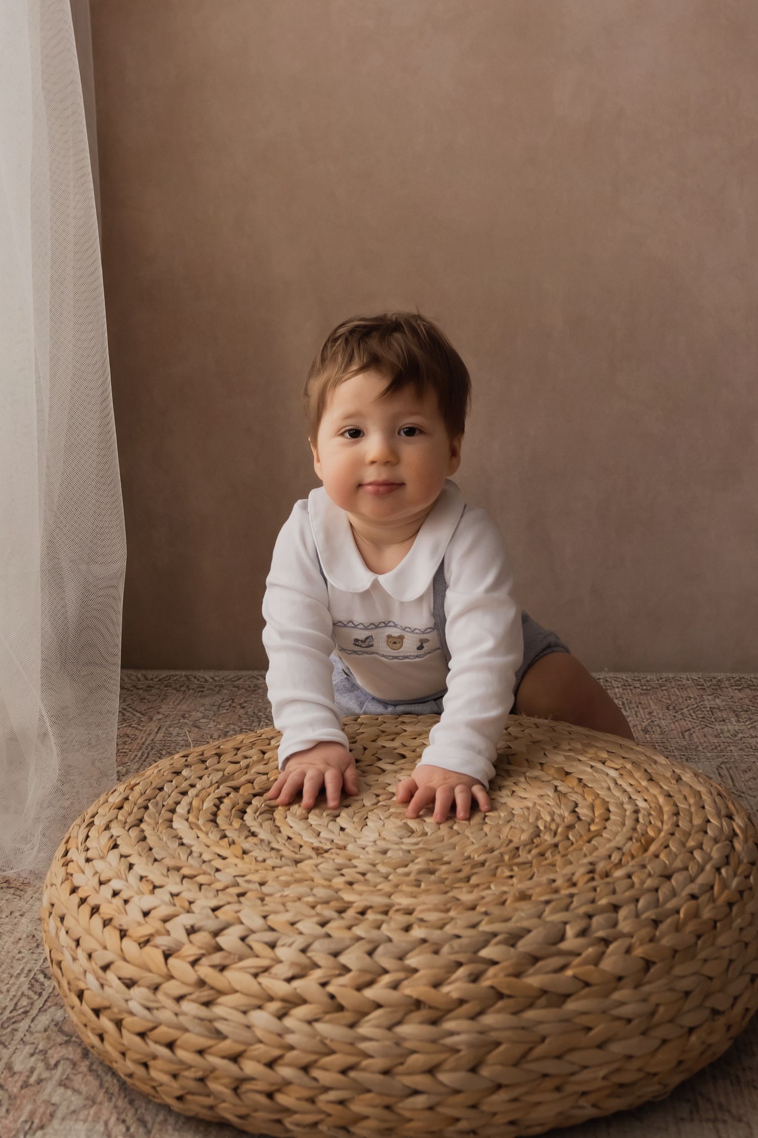 Curious baby boy in white onesie leaning on woven rattan pouf on patterned rug.