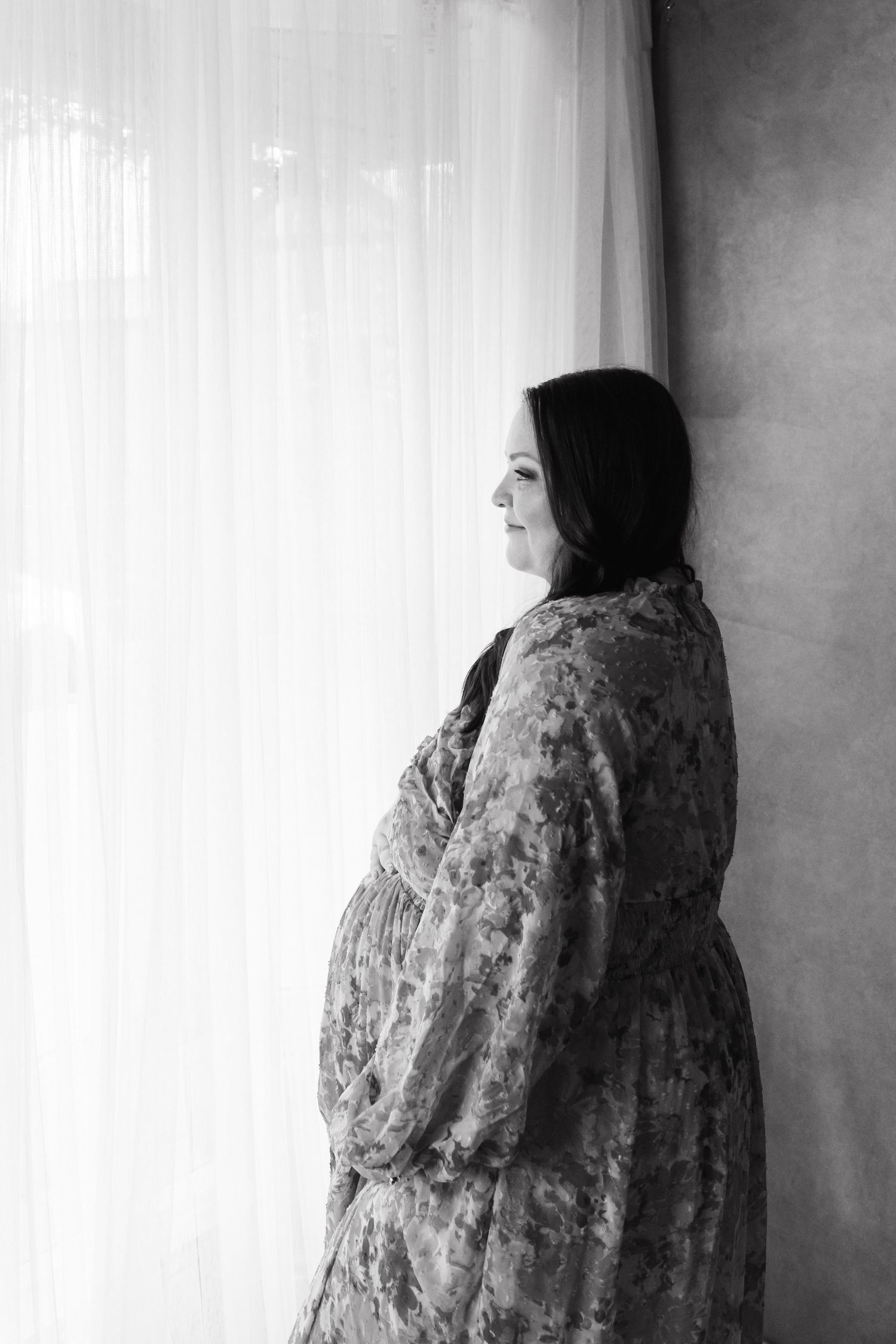 Black and white portrait of a pregnant woman in floral robe gazing out a window.