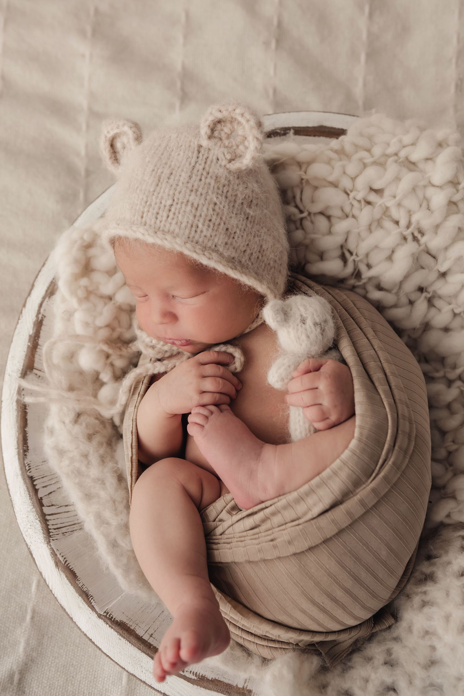 Sleeping newborn baby in beige bear ear bonnet hat, wrapped in cream fabric inside a white wooden bowl.