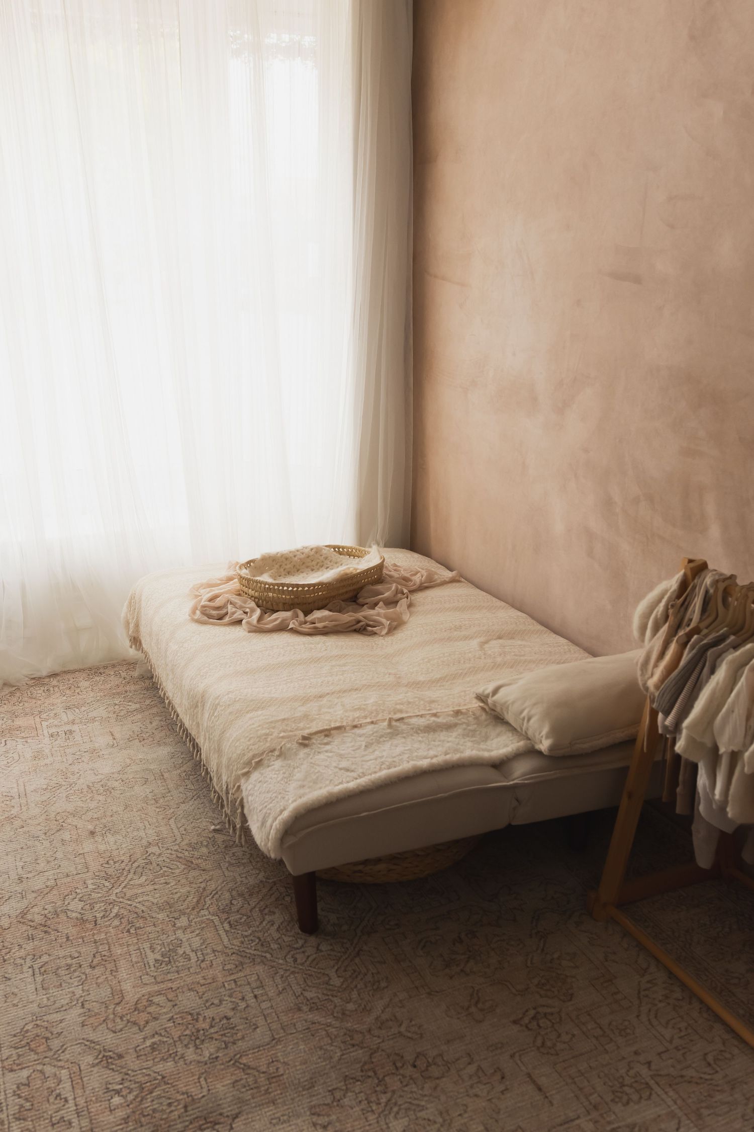 Minimalist boho bedroom with a linen daybed, straw hat, sheer curtains, and warm beige textured walls on a vintage rug.