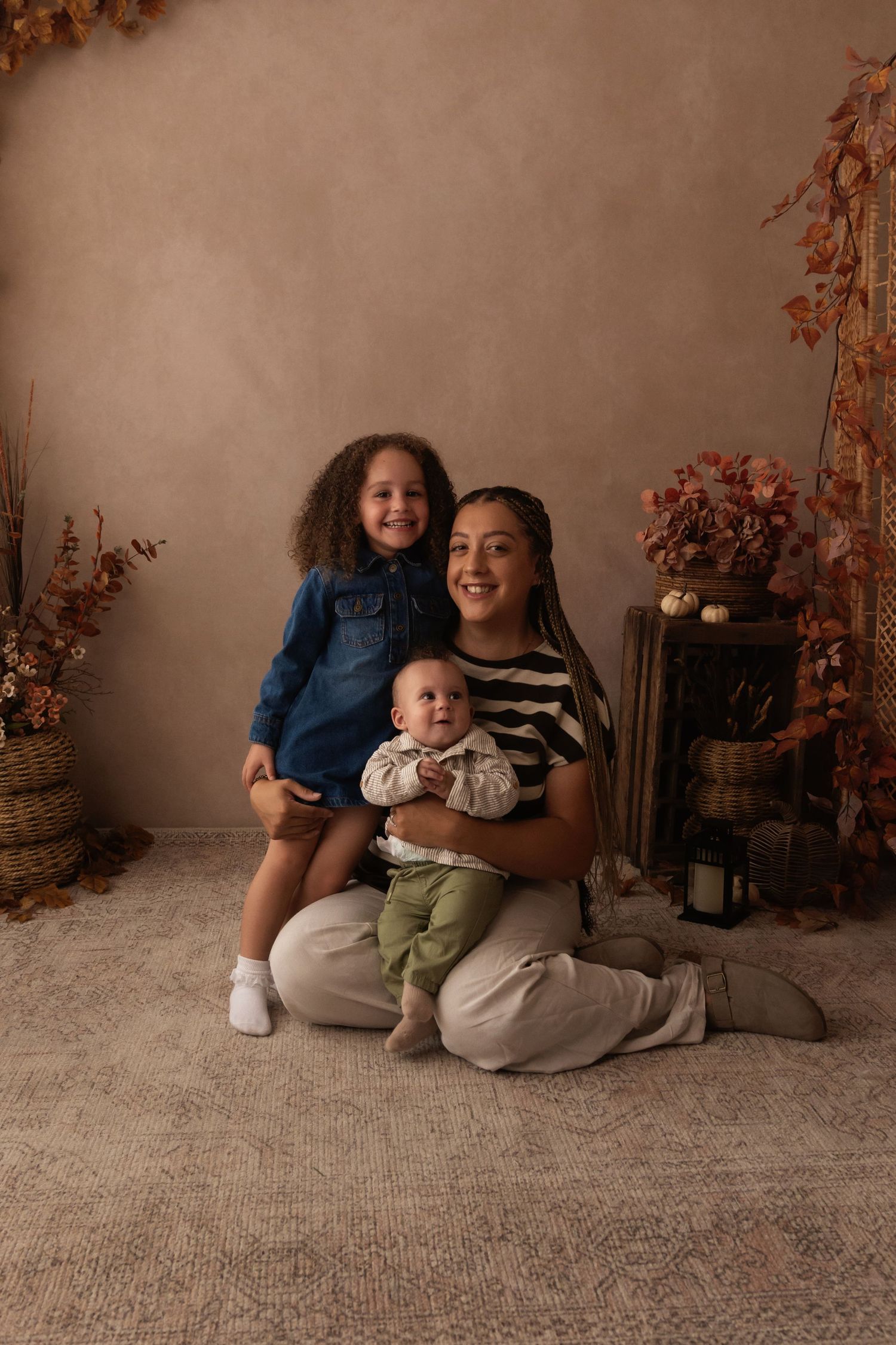 A mother sits on a carpet holding her baby and young daughter in a warm fall-themed studio portrait setting.