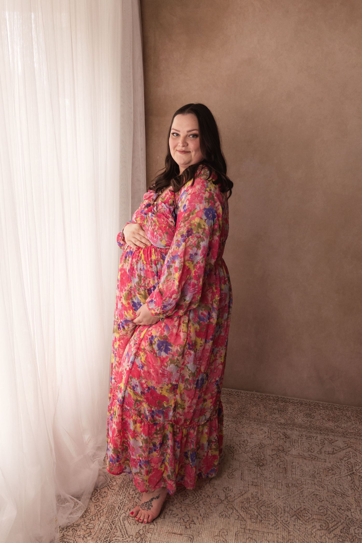 A pregnant woman in a vibrant floral pink maxi dress poses gracefully by a sheer curtain.