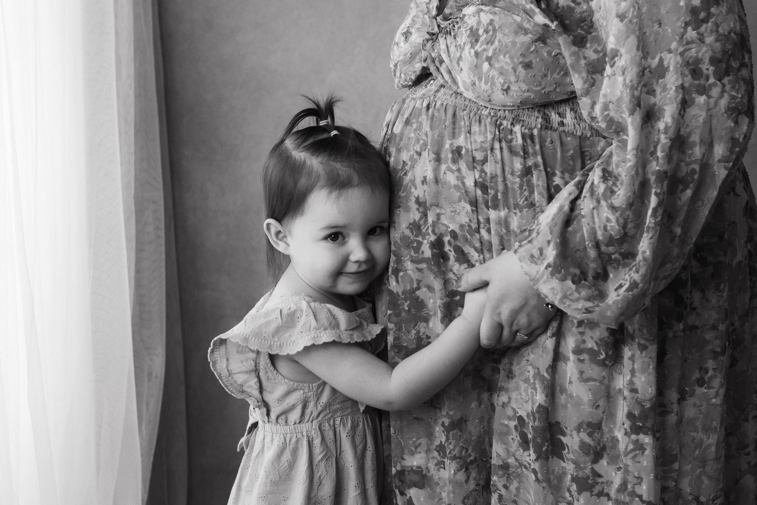 Black and white photo of a toddler girl in a dress smiling while touching her pregnant mother's gown.
