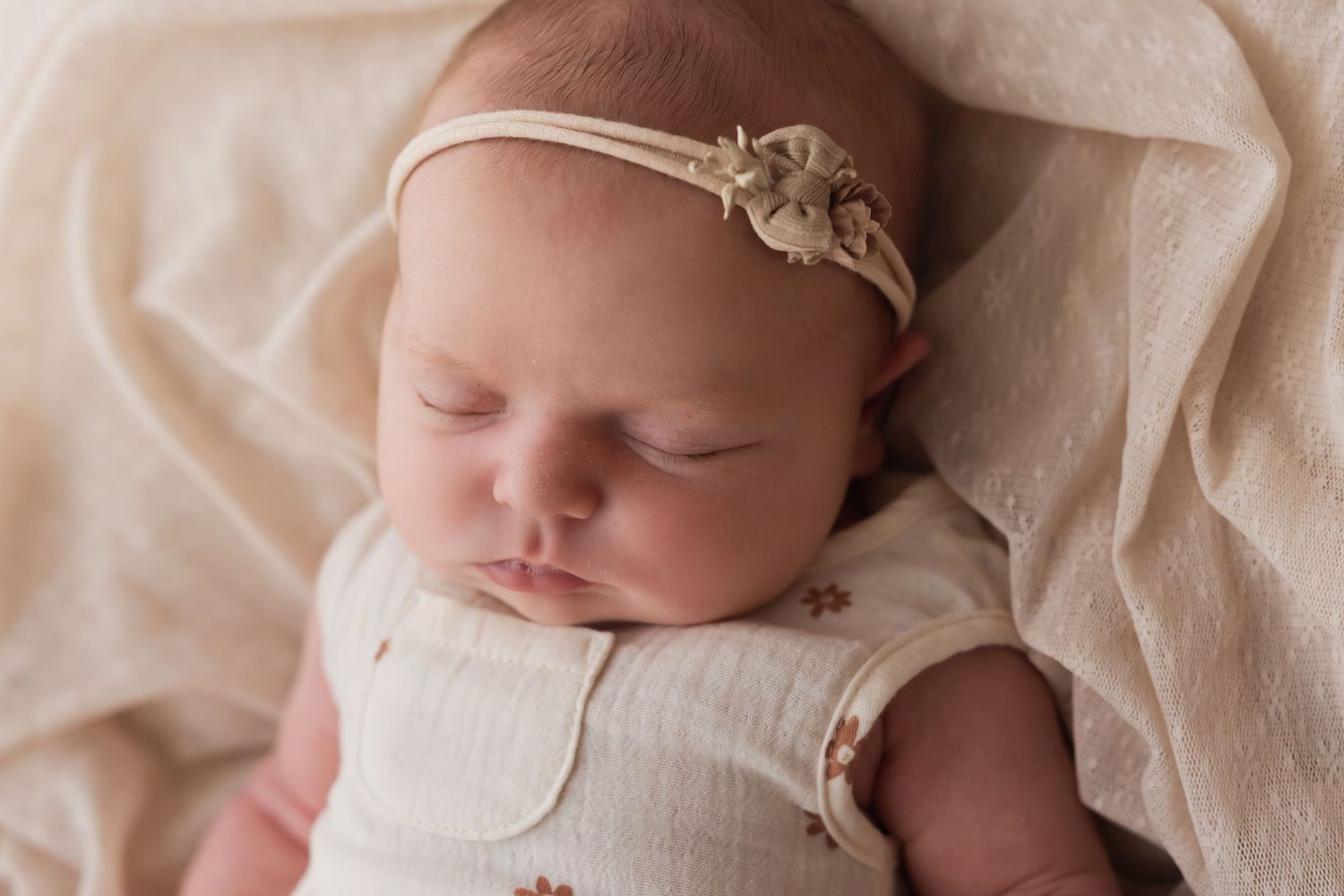 Peaceful newborn baby girl sleeping in cream floral onesie and delicate flower headband on soft beige blanket.